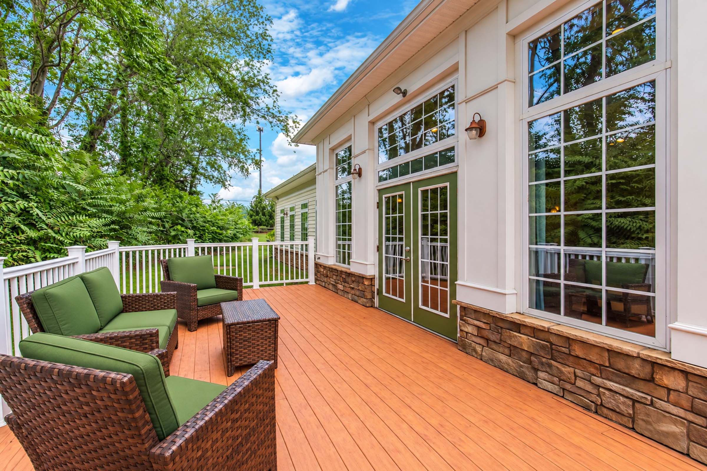 A spacious outdoor deck featuring green armchairs and a small table, surrounded by lush greenery and large windows of a modern building. The wooden flooring complements the stone accents and white railings, creating an inviting and relaxed atmosphere.