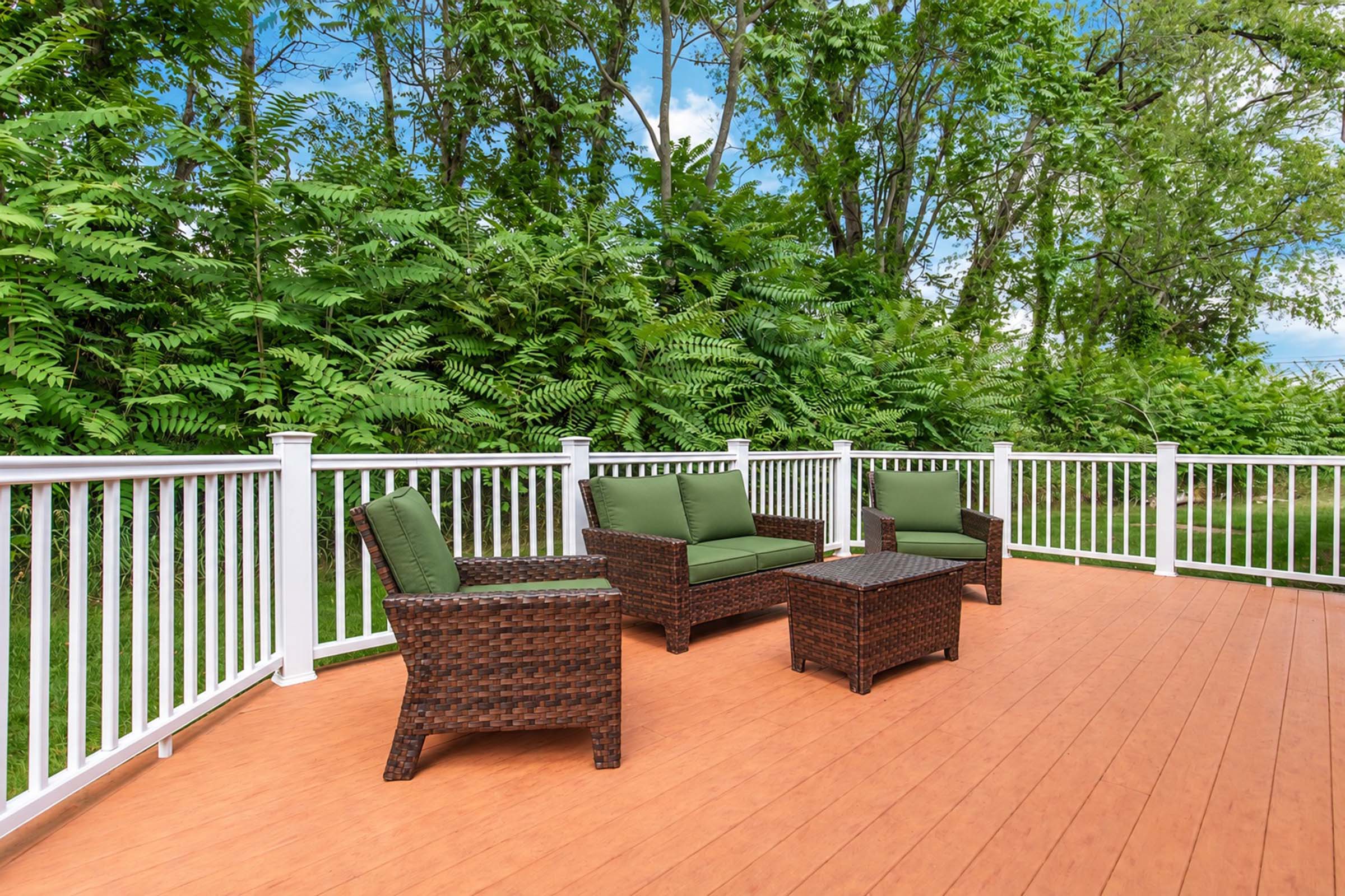 A cozy outdoor patio featuring brown wicker furniture with green cushions, including a loveseat, two chairs, and a small table. The patio is surrounded by lush greenery and a white railing, with a wooden deck beneath. The scene evokes a serene and inviting atmosphere for relaxation.