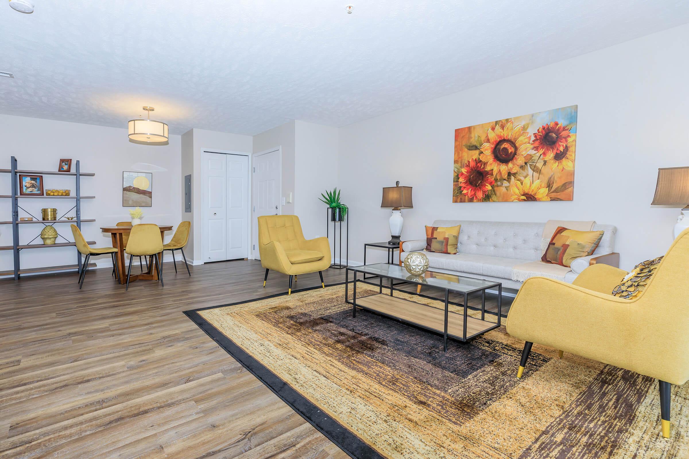 A bright and spacious living area featuring a comfortable gray sofa with yellow accent chairs. A decorative coffee table sits on a textured rug, and a colorful sunflower artwork adorns the wall. In the background, a dining table with yellow chairs is visible, along with a shelf displaying decorative items.