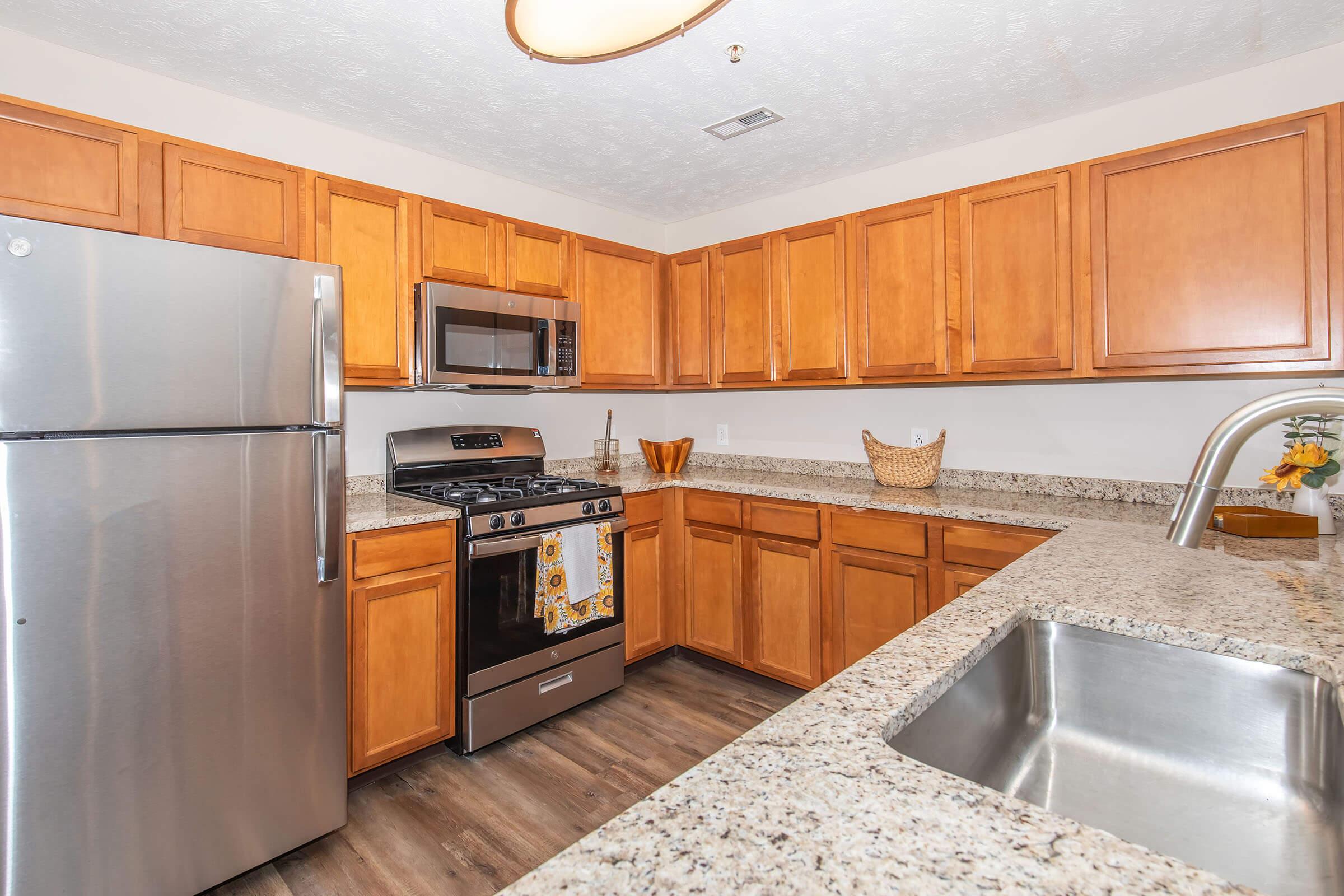 A modern kitchen featuring wooden cabinets, stainless steel appliances including a refrigerator and gas stove, a microwave, and a granite countertop. A stainless steel sink is visible, along with a basket and decorative items on the counter. The kitchen has warm lighting and a spacious layout.