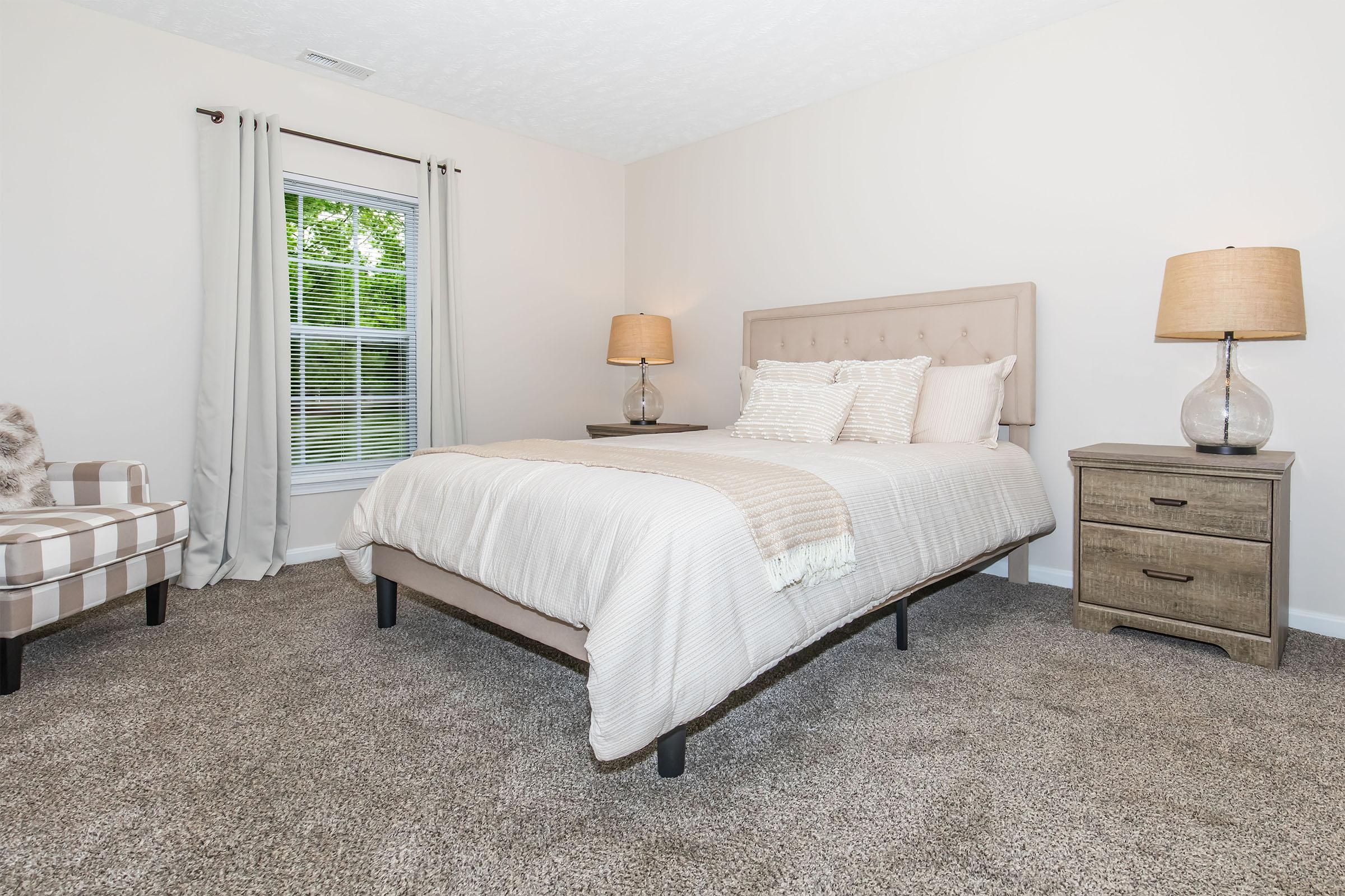 A cozy bedroom featuring a plush bed with light-colored bedding, two bedside lamps, and a wooden nightstand. A comfortable armchair with a checkered pattern is placed in the corner. Natural light filters through a window covered with sheer curtains, illuminating the neutral-toned walls and carpet.
