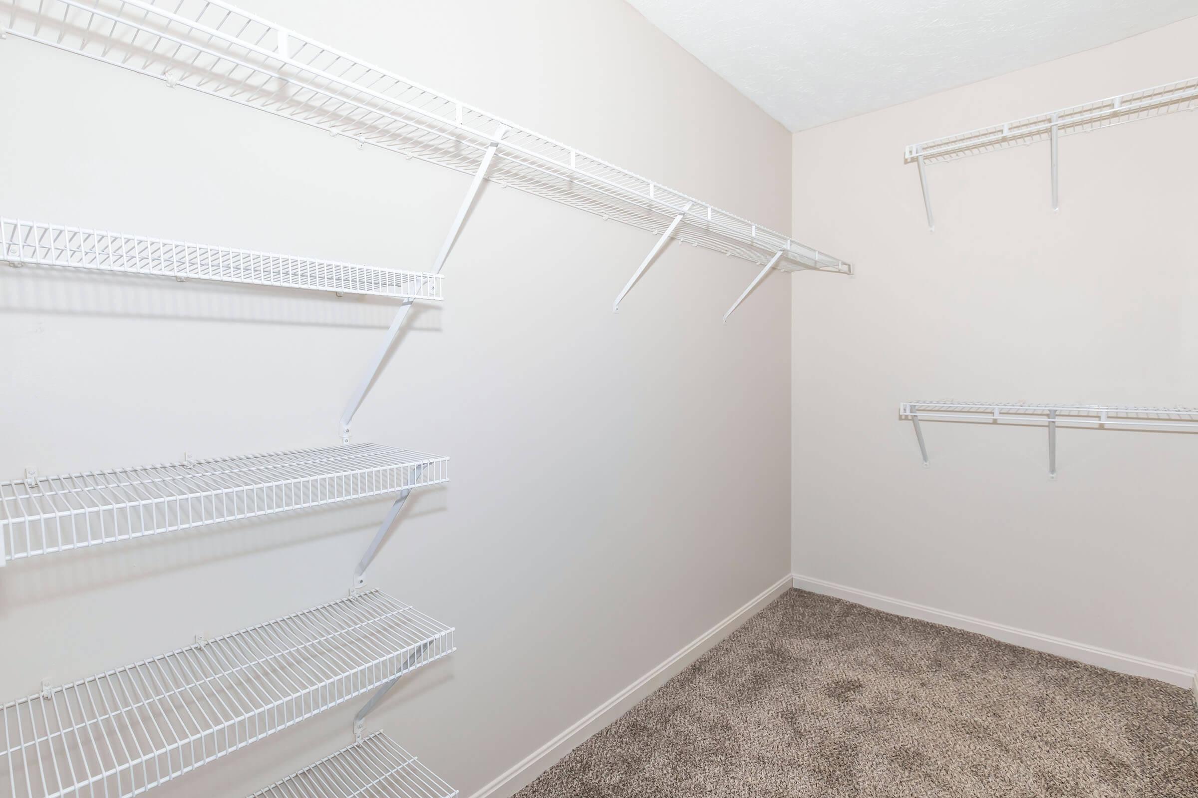 A neatly organized empty closet featuring white wire shelving on beige walls. The floor is covered with tan carpet, and the shelves are positioned for optimal storage, providing ample blank space for hanging and storing items.