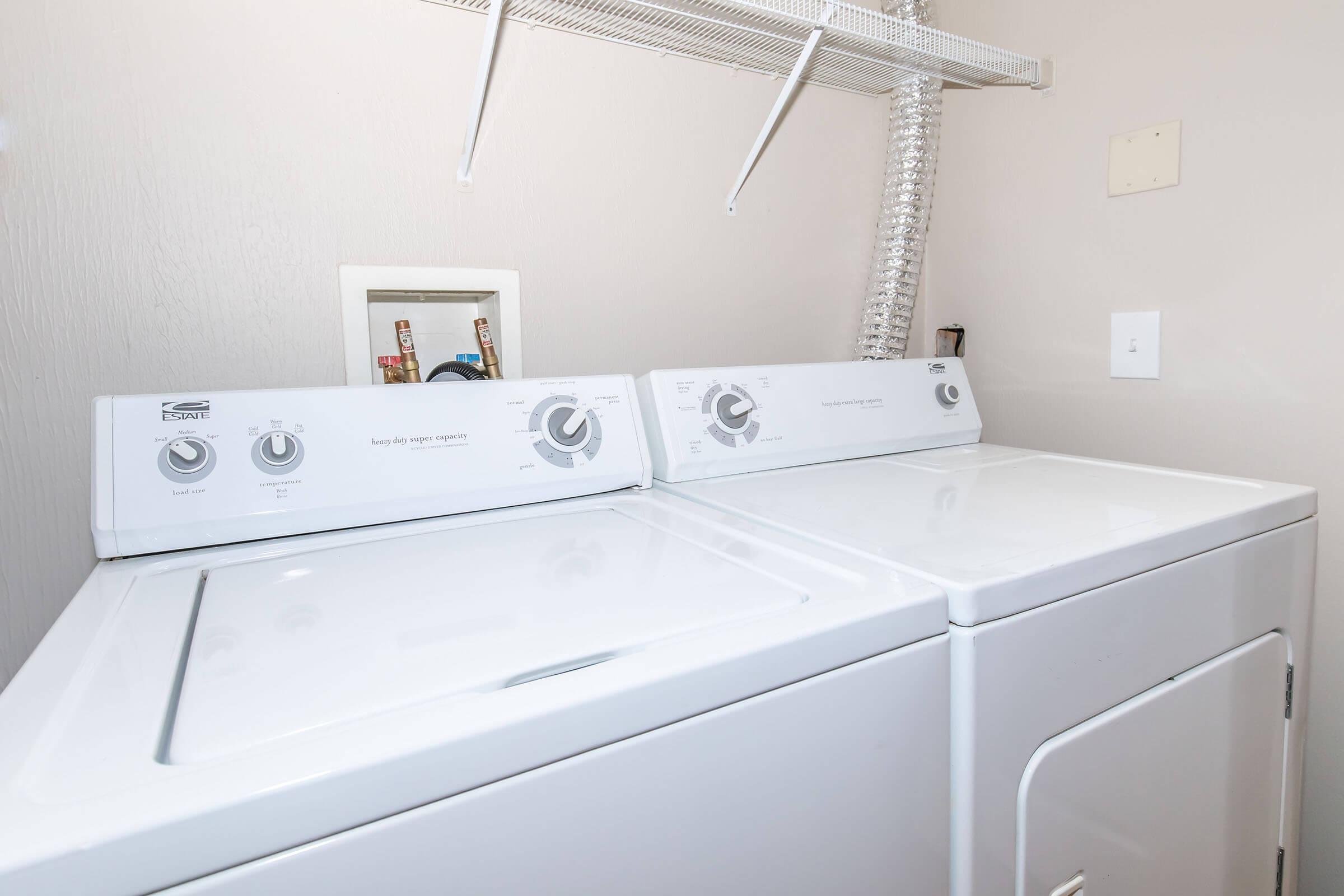 A laundry room featuring a white washer and dryer set side by side, with control dials visible. The wall behind is light-colored, and there's a vent pipe above the appliances. A power outlet is visible on the right wall, adding to the functional, clean ambiance of the space.