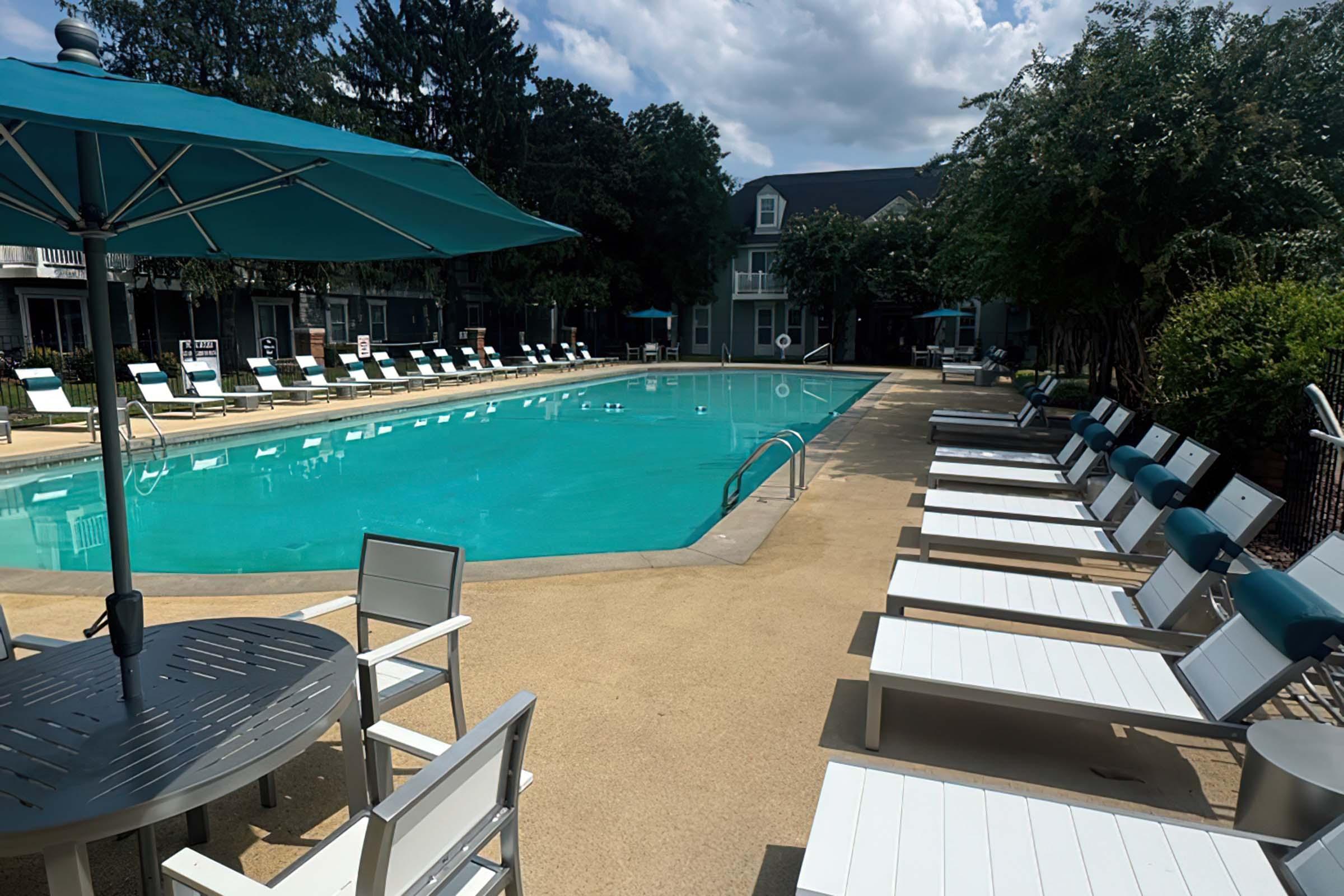 A tranquil outdoor pool area featuring a clear blue swimming pool surrounded by lounge chairs and tables. A large teal umbrella provides shade. Lush greenery and buildings in the background create a relaxing atmosphere on a sunny day.