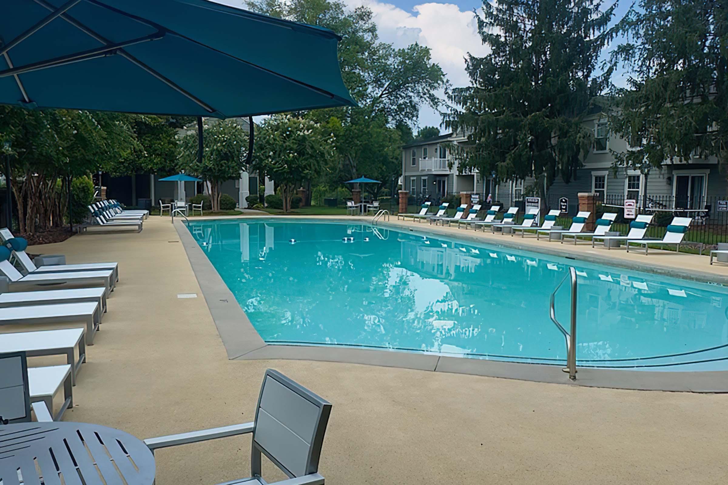 A clear swimming pool surrounded by lounge chairs and umbrellas, set in a landscaped area with trees. Nearby buildings feature a neutral color palette. The sky is partly cloudy, and the pool area appears well-maintained and inviting.