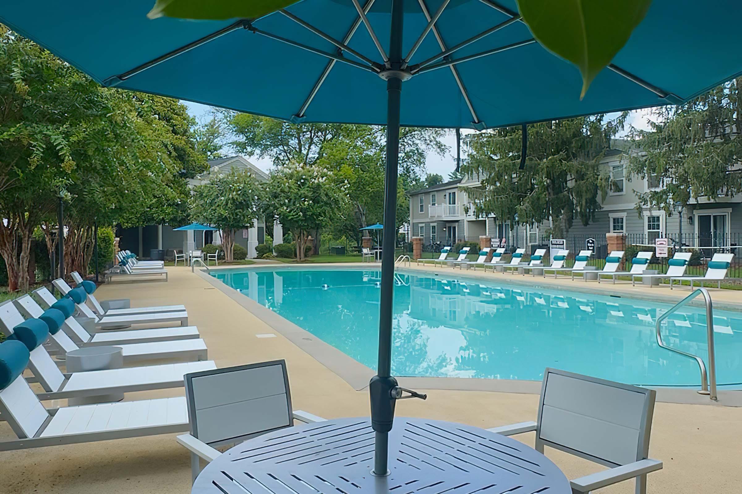 A view of a swimming pool area featuring a clear blue pool, surrounded by lounge chairs and umbrellas. In the foreground, a table with an umbrella provides shade, with trees and residential buildings visible in the background. The scene is peaceful and inviting, ideal for relaxation.
