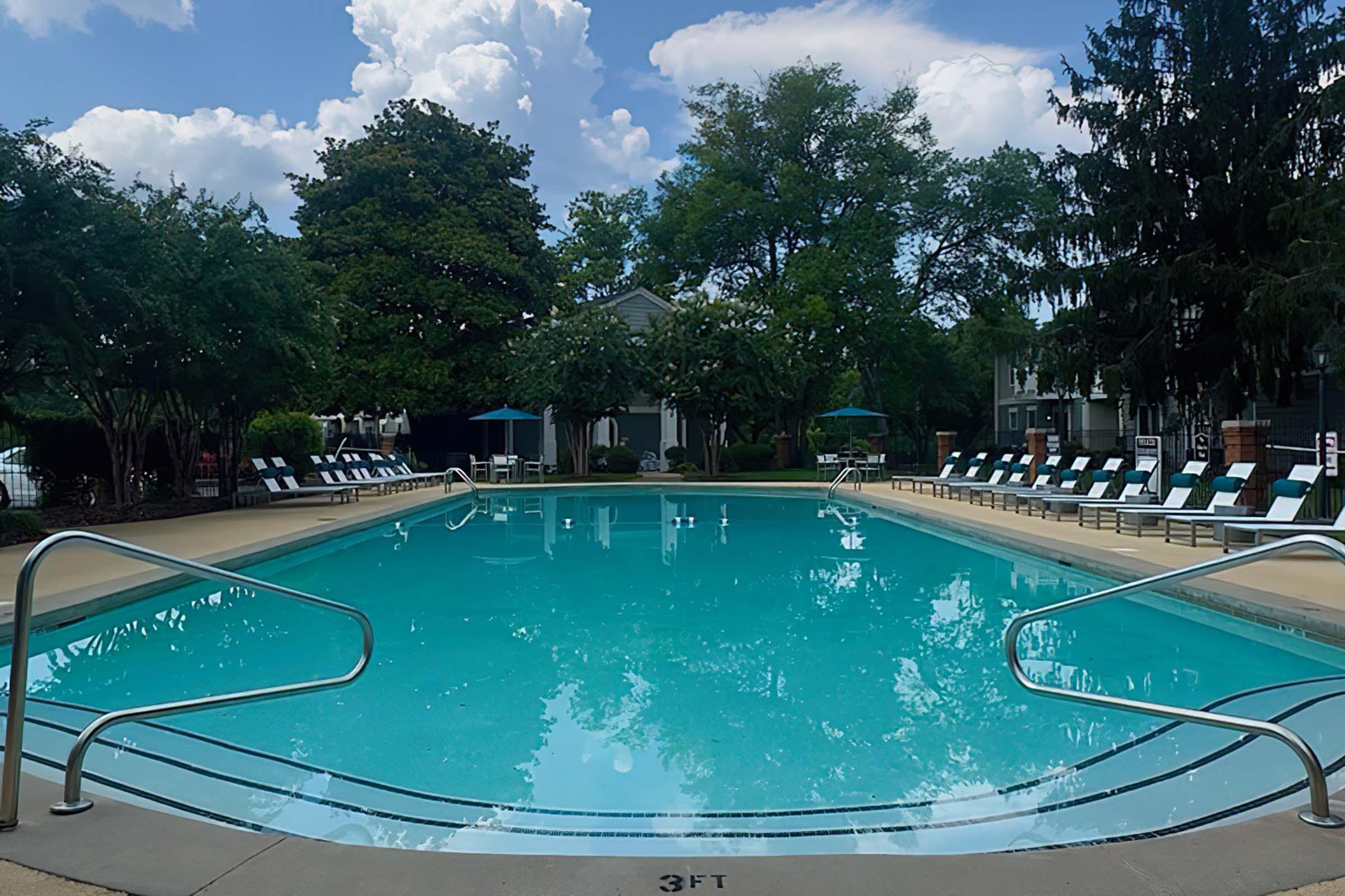 A clean, inviting swimming pool surrounded by lush greenery and sun loungers. The pool features a deep end and a shallow end marked with "3 FT." Fluffy white clouds drift in a blue sky above, creating a relaxing atmosphere. Sunshades are set up around the pool area, ready for guests to enjoy.
