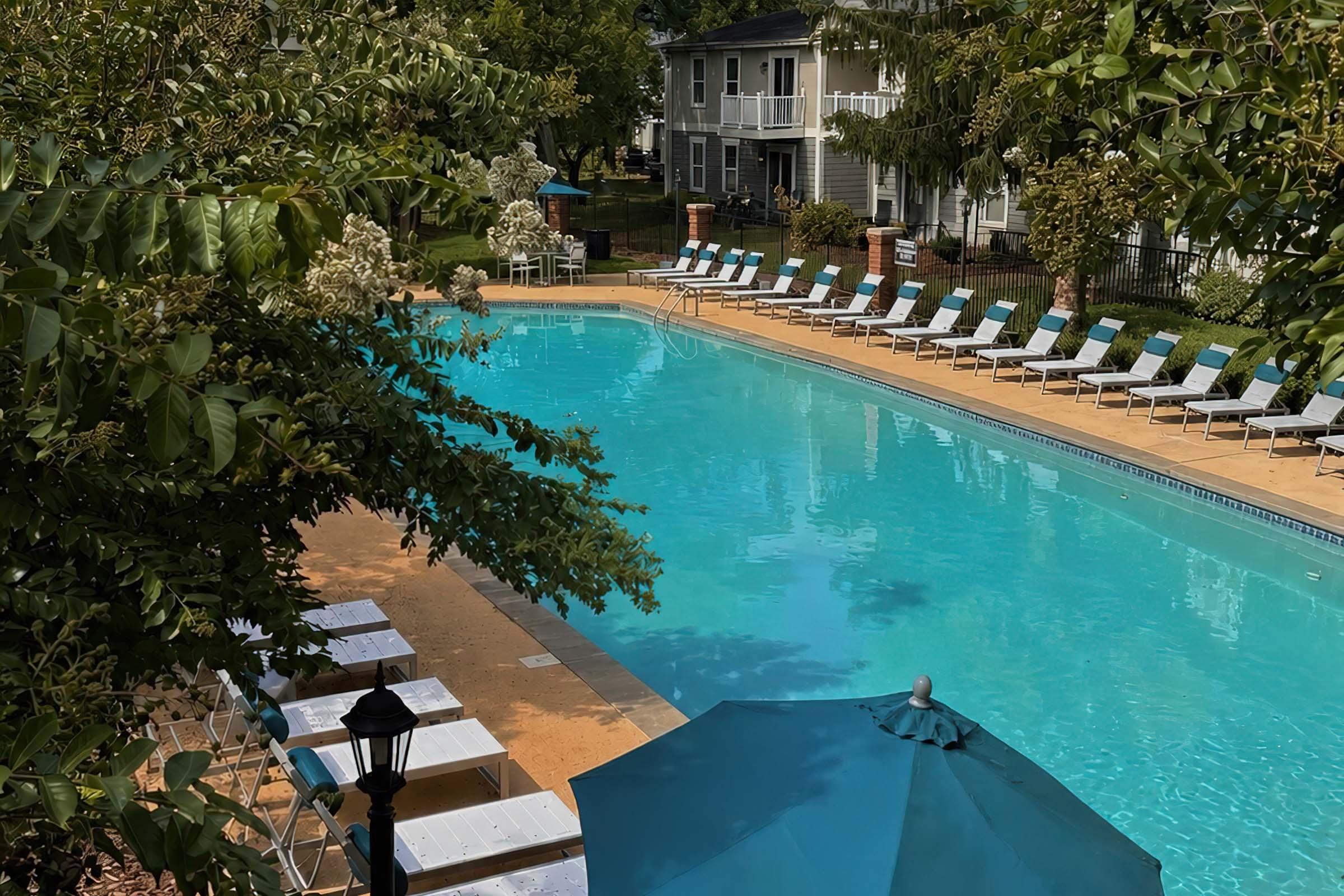 A serene outdoor pool area surrounded by lush trees. The turquoise water reflects sunlight, with several lounge chairs arranged neatly along the poolside. An umbrella provides shade for a couple of seating areas. In the background, there are multi-story residential buildings, creating a tranquil and inviting atmosphere.