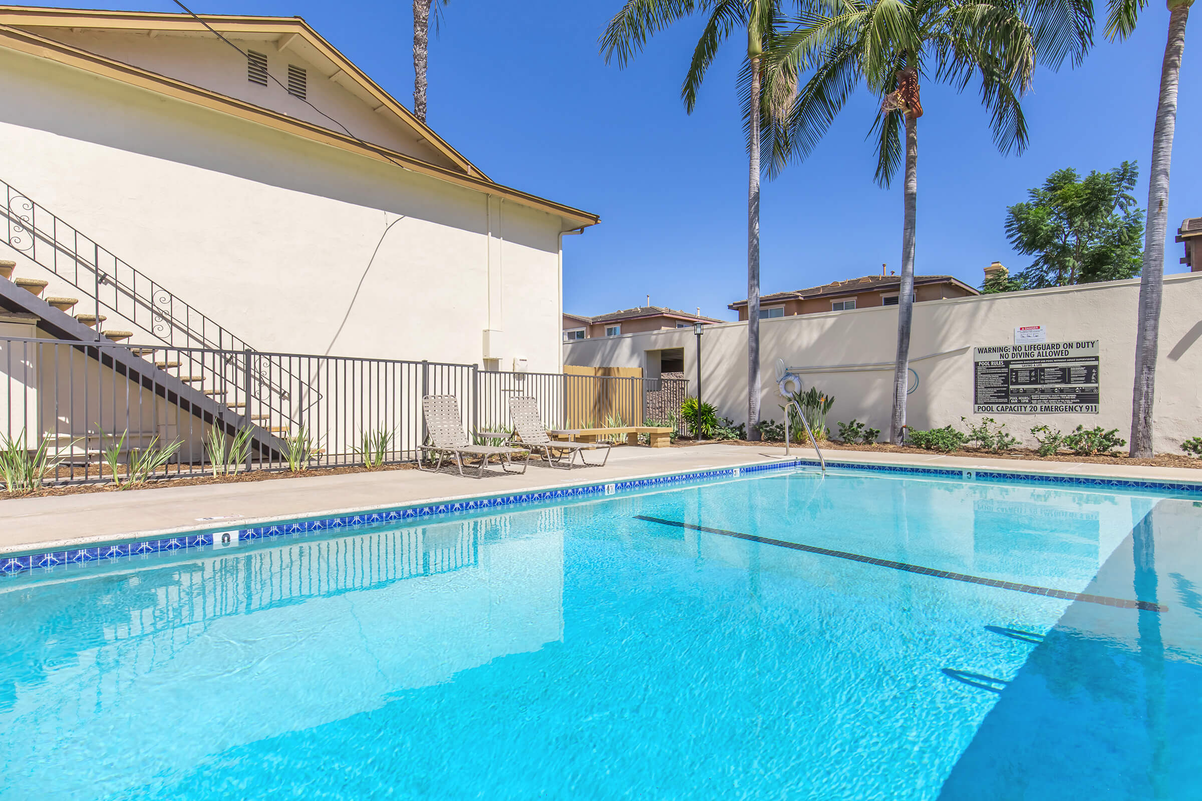 A clear blue swimming pool surrounded by lounge chairs and palm trees, with a white building in the background. The scene is sunny and inviting, showcasing a well-maintained outdoor area perfect for relaxation and leisure.