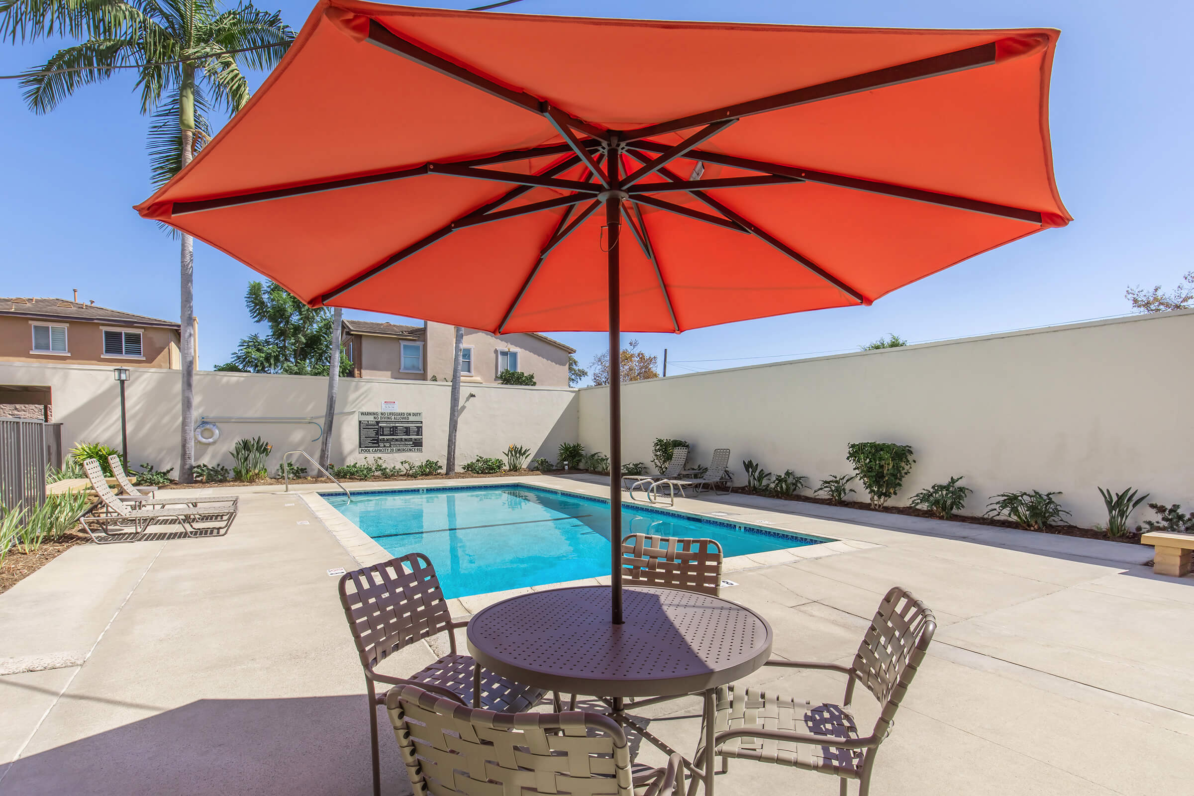A bright orange umbrella shades a table and chairs by a swimming pool. The pool is surrounded by a clean patio with lush greenery and palm trees in the background. Sunlight fills the area, creating a warm and inviting atmosphere for relaxation.