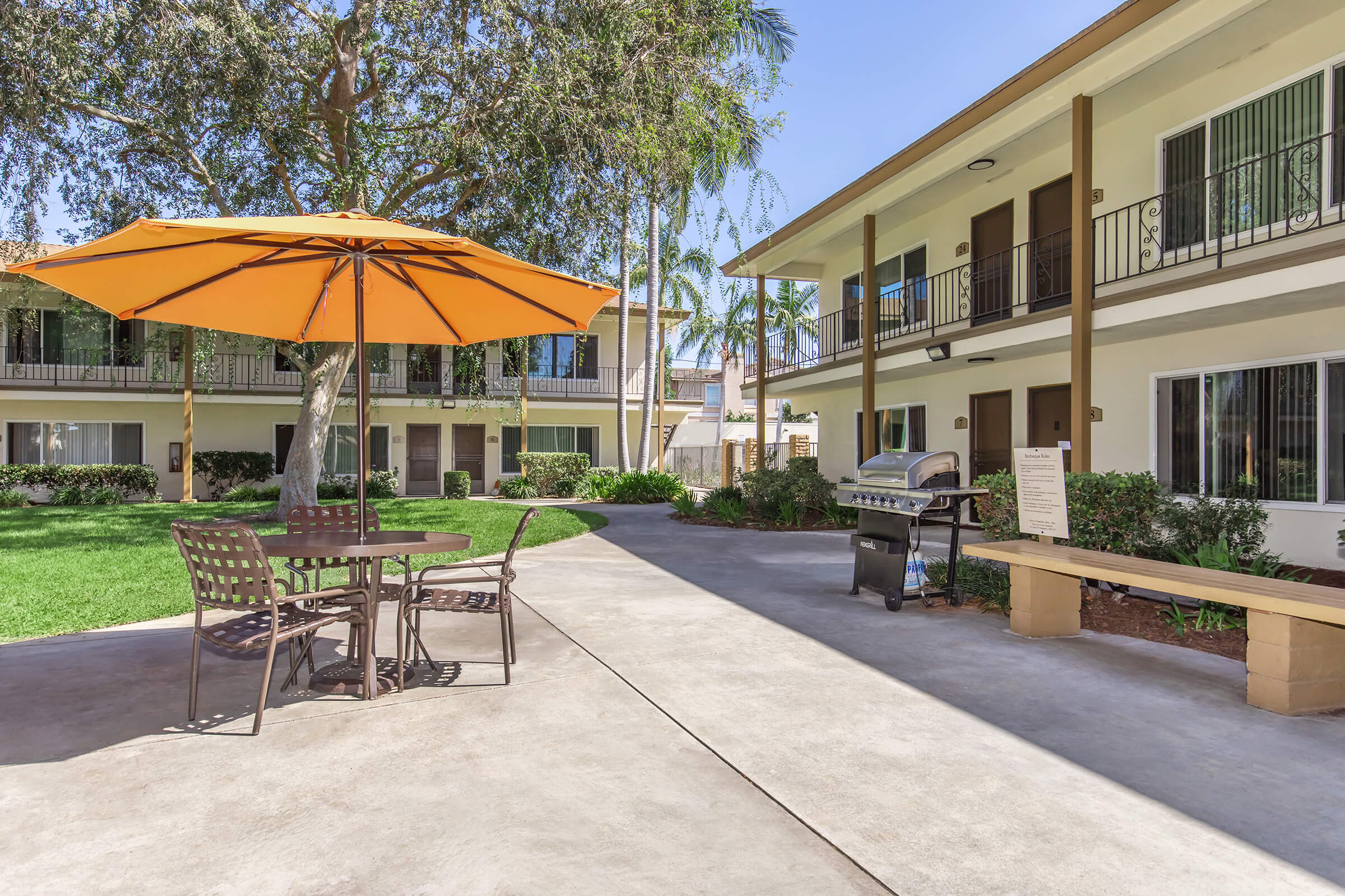 A well-maintained outdoor area featuring a round table with four chairs under a large orange umbrella. Nearby, there's a barbecue grill and landscaped greenery. The background includes two beige apartment buildings with balconies and large windows, set in a sunny environment with palm trees.