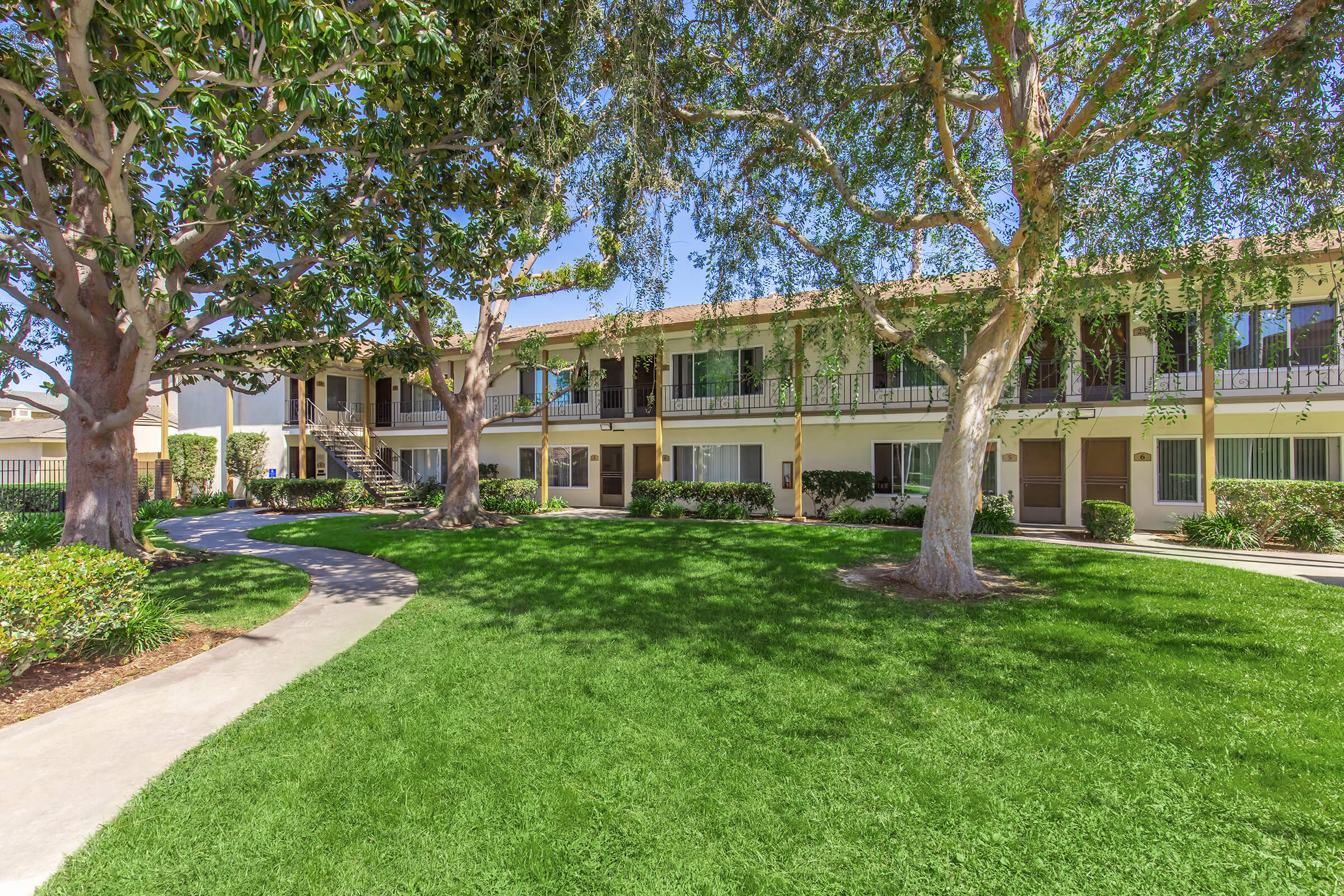 A well-maintained apartment complex with two-story buildings surrounded by lush green lawns and trees. A curved pathway leads through the grassy area, providing access to the individual units. The scene is bright and inviting, suggesting a peaceful residential environment.