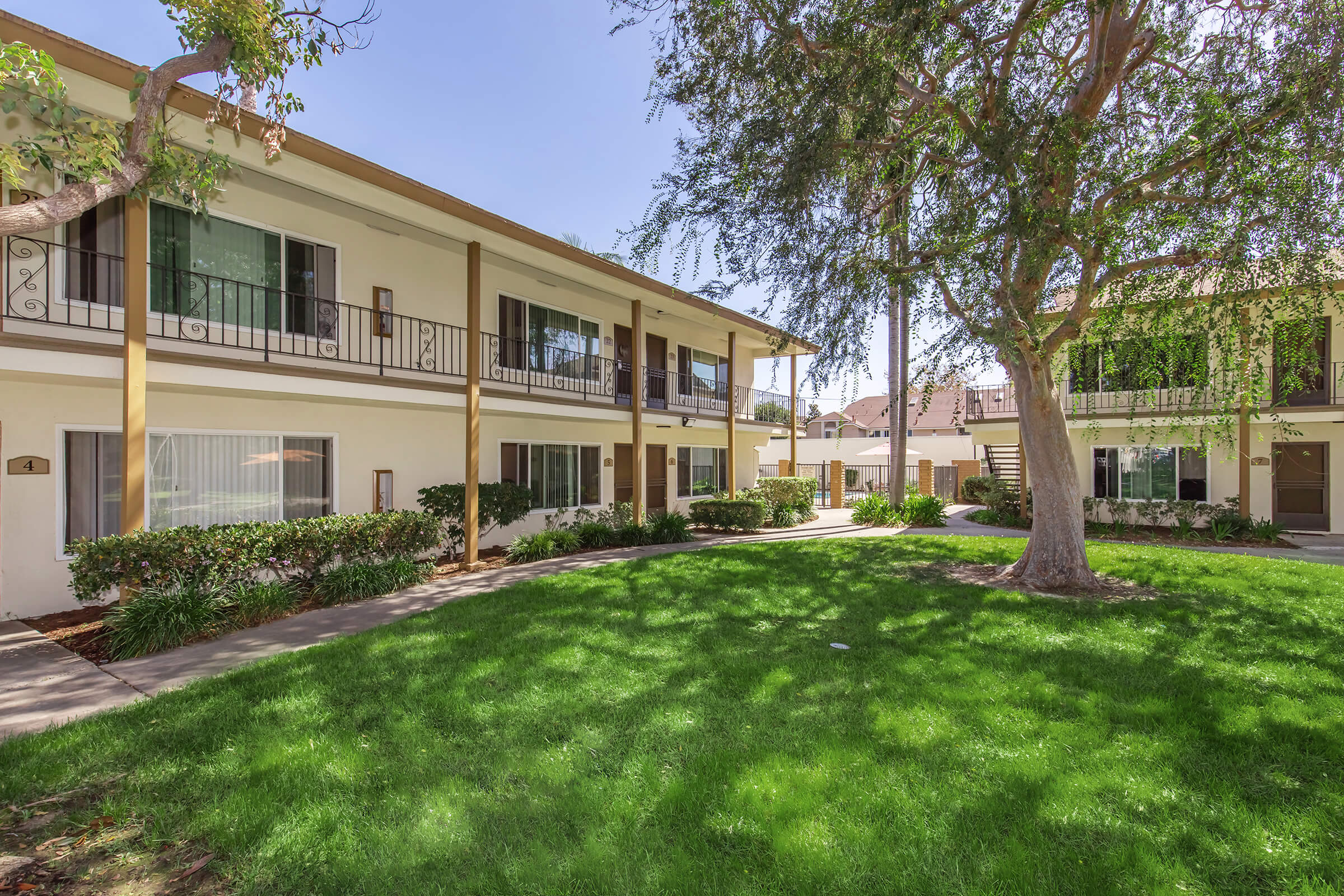 A landscaped courtyard with lush green grass and trees, surrounded by two-story residential buildings. The buildings feature balconies and windows, creating a peaceful, inviting atmosphere in a sunny environment. Pathways lead through the greenery, enhancing the residential appeal.