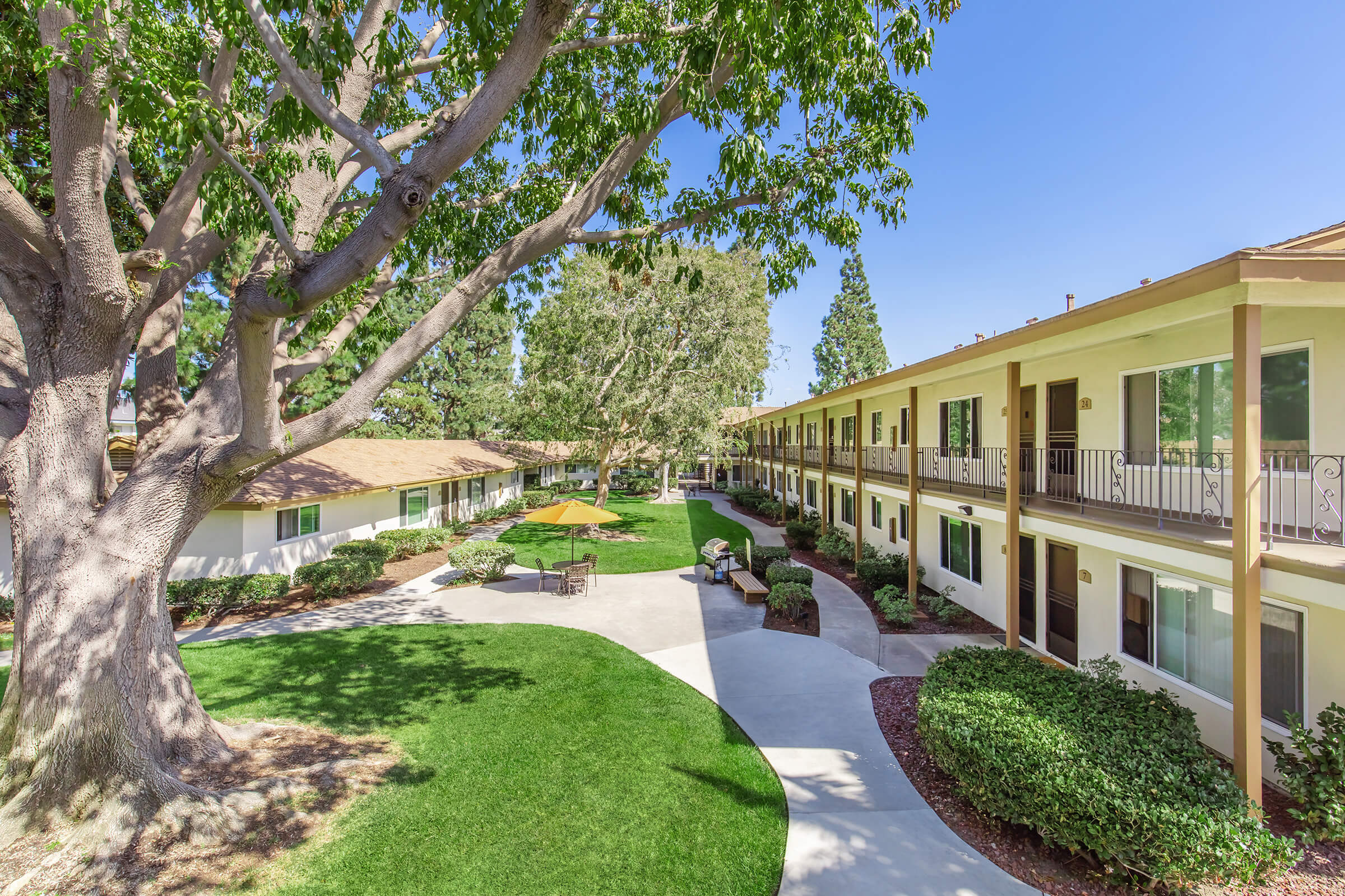 A view of a well-maintained courtyard with lush green grass and trees. Surrounding the courtyard are two-story buildings with balconies. In the center, there are outdoor seating areas and a yellow umbrella, creating a relaxing atmosphere. The sky is clear and blue, adding to the pleasant environment.