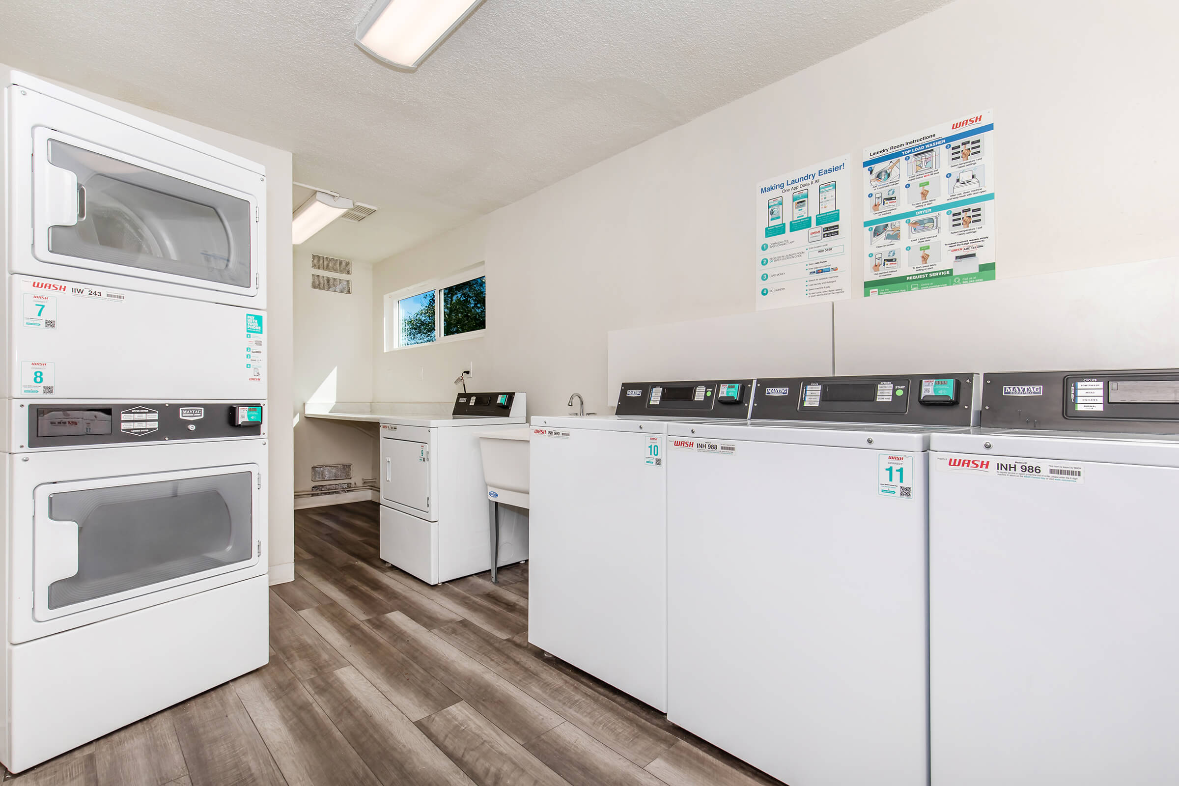 A clean and well-lit laundry room featuring multiple stacked washing machines and dryers. There's a folding space and a sink on one side, with informational posters on the wall providing instructions and guidelines for using the machines. The flooring is modern and the walls are painted a neutral color.
