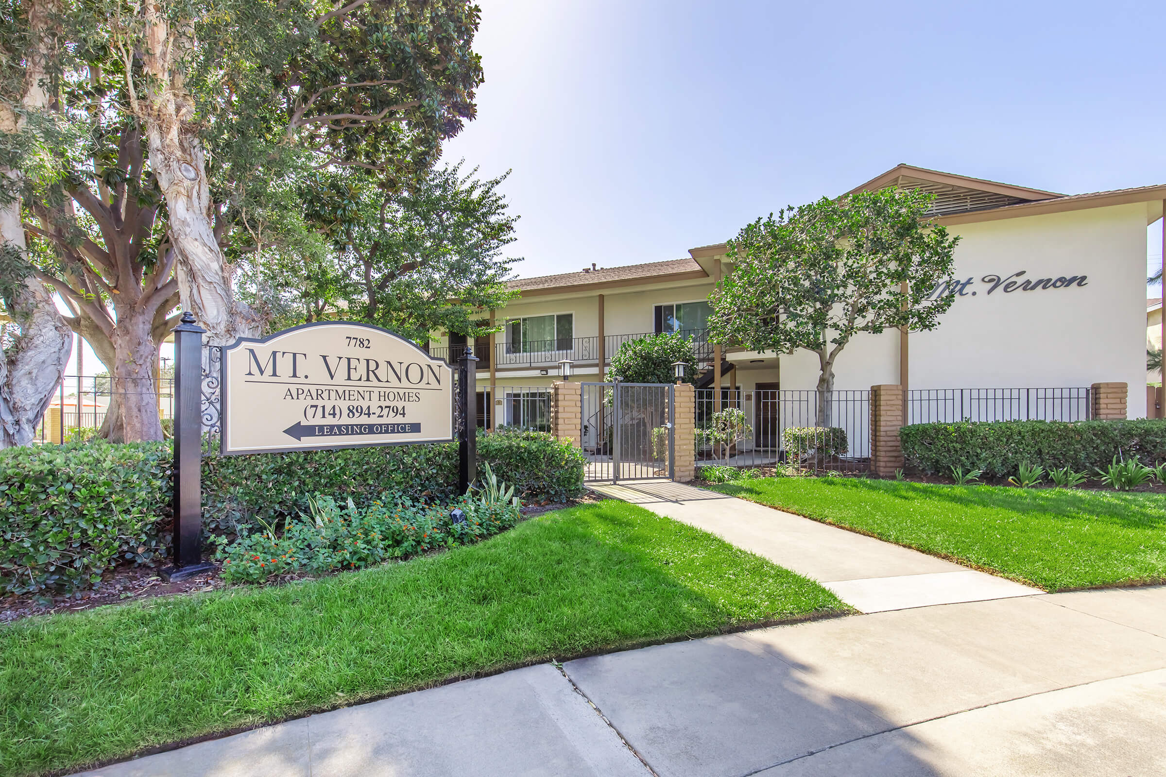 Sign for "Mt. Vernon Apartment Homes" at 7792 with contact number (714) 894-2704, surrounded by green landscaping and trees. A pathway leads to the entrance of the apartment complex, which features a two-story building in the background.