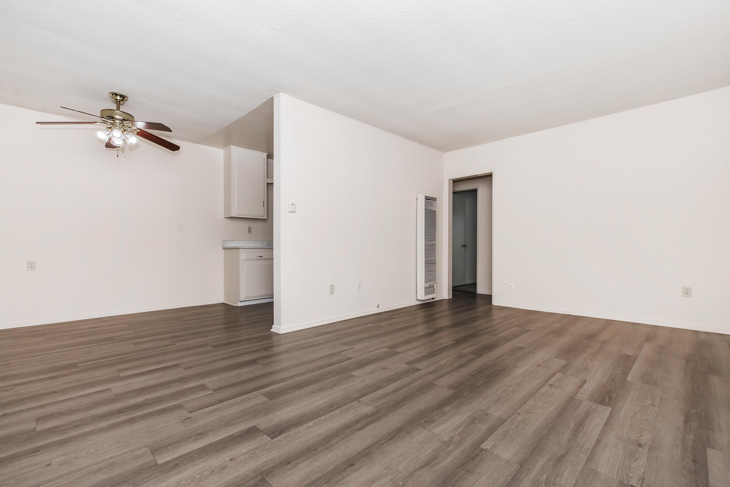 A spacious, empty living area with light-colored walls and a smooth, wooden floor. A ceiling fan is visible in the corner, and a small kitchenette is partially visible in the background. The room is well-lit, with doorways leading to other areas, creating an open and airy atmosphere.