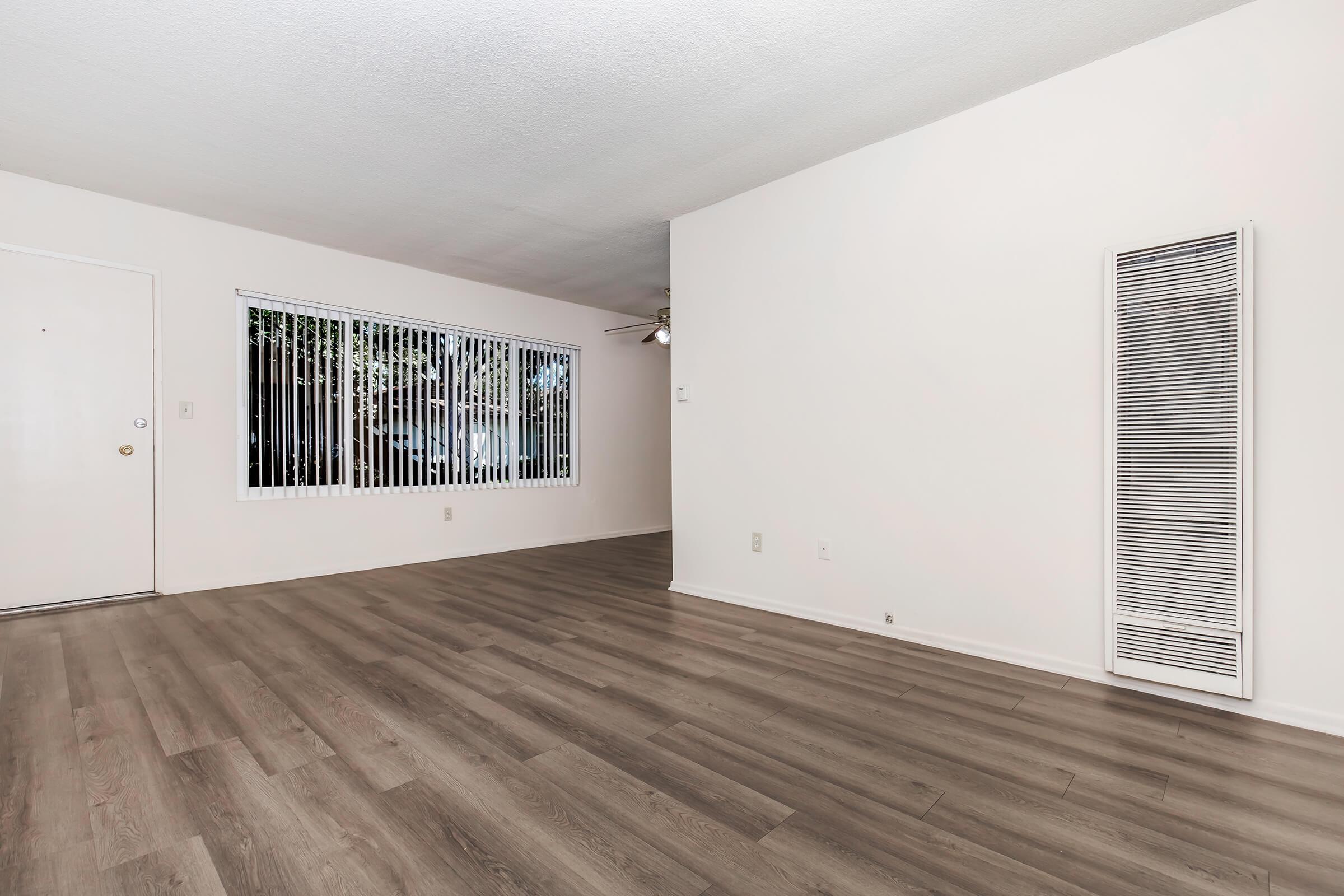 An empty room with light-colored walls and a large window featuring white blinds. The floor is covered in brown laminate, and there is a wall-mounted heater on the right side. A ceiling fan is visible above, and the space is well-lit, creating a bright and open atmosphere.