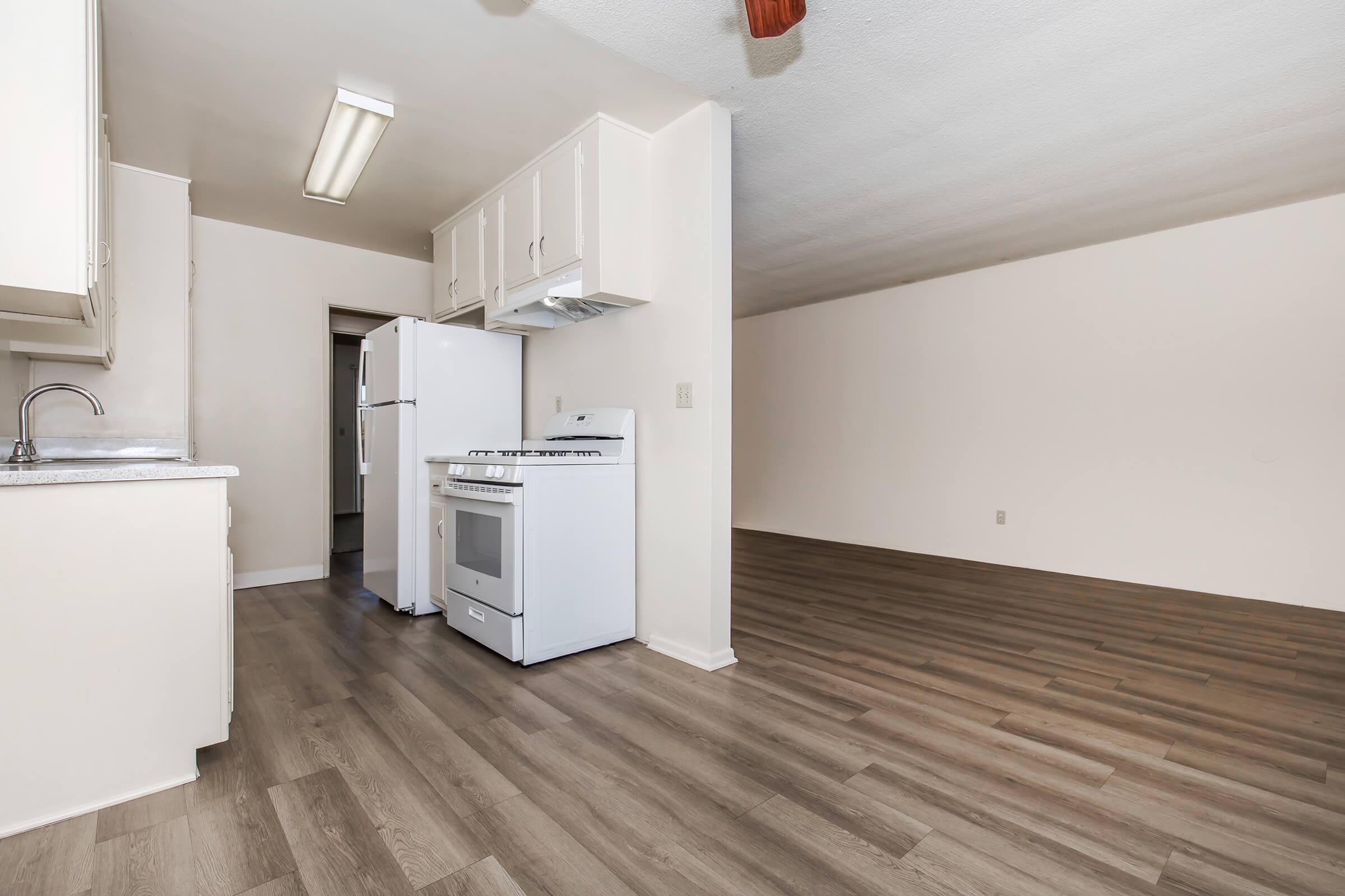 A bright kitchen area featuring white cabinets, a white refrigerator, and a gas stove. The flooring is light-colored laminate, creating a spacious feel. In the background, there is an open area with plain walls and a ceiling fan, suggesting a seamless transition between the kitchen and living space.