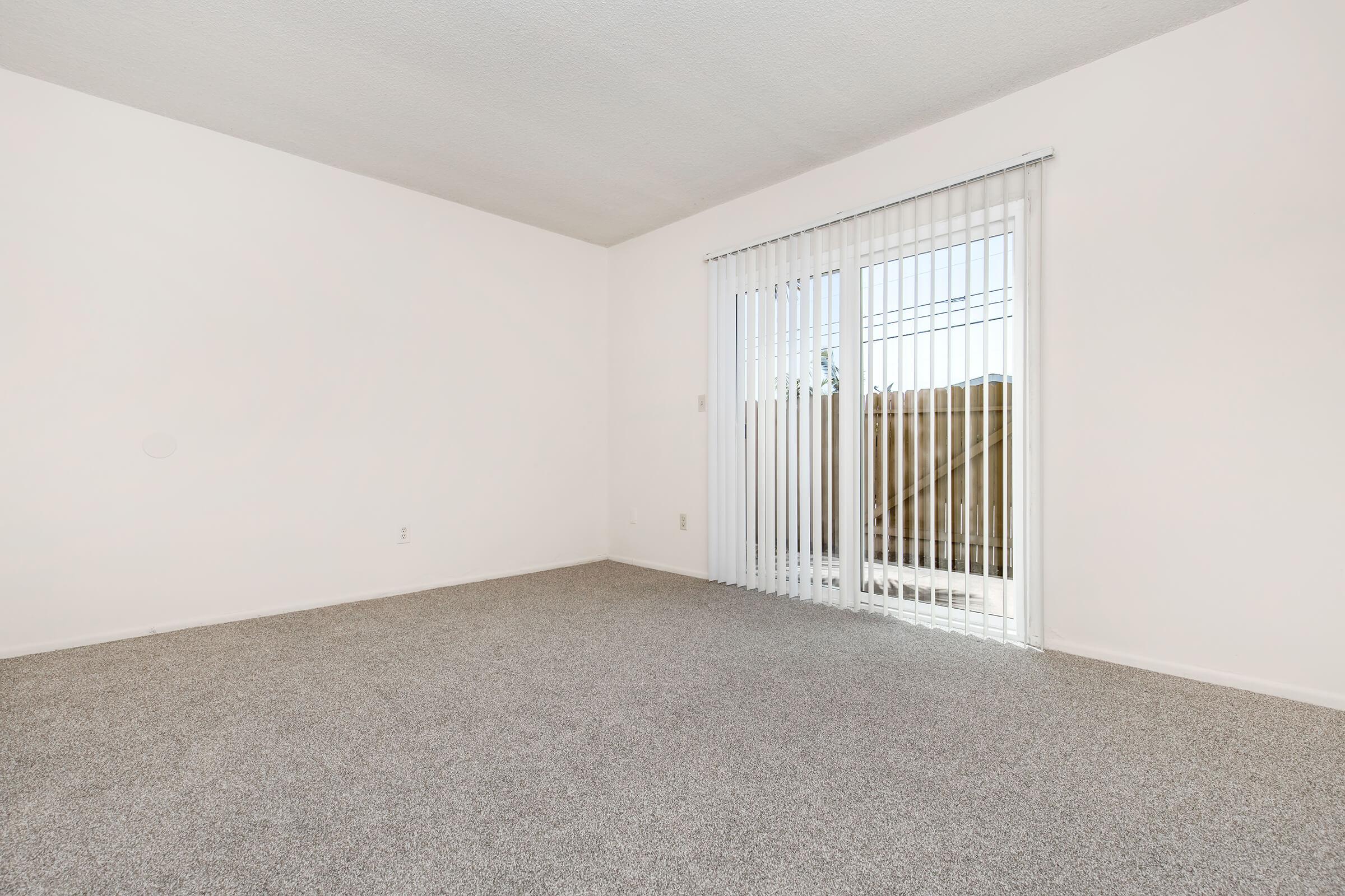 A vacant room with light gray carpet and plain white walls. There is a set of sliding glass doors with vertical blinds leading to an outdoor space. The room appears bright and airy, with a minimalist design and ample natural light coming through the doors.
