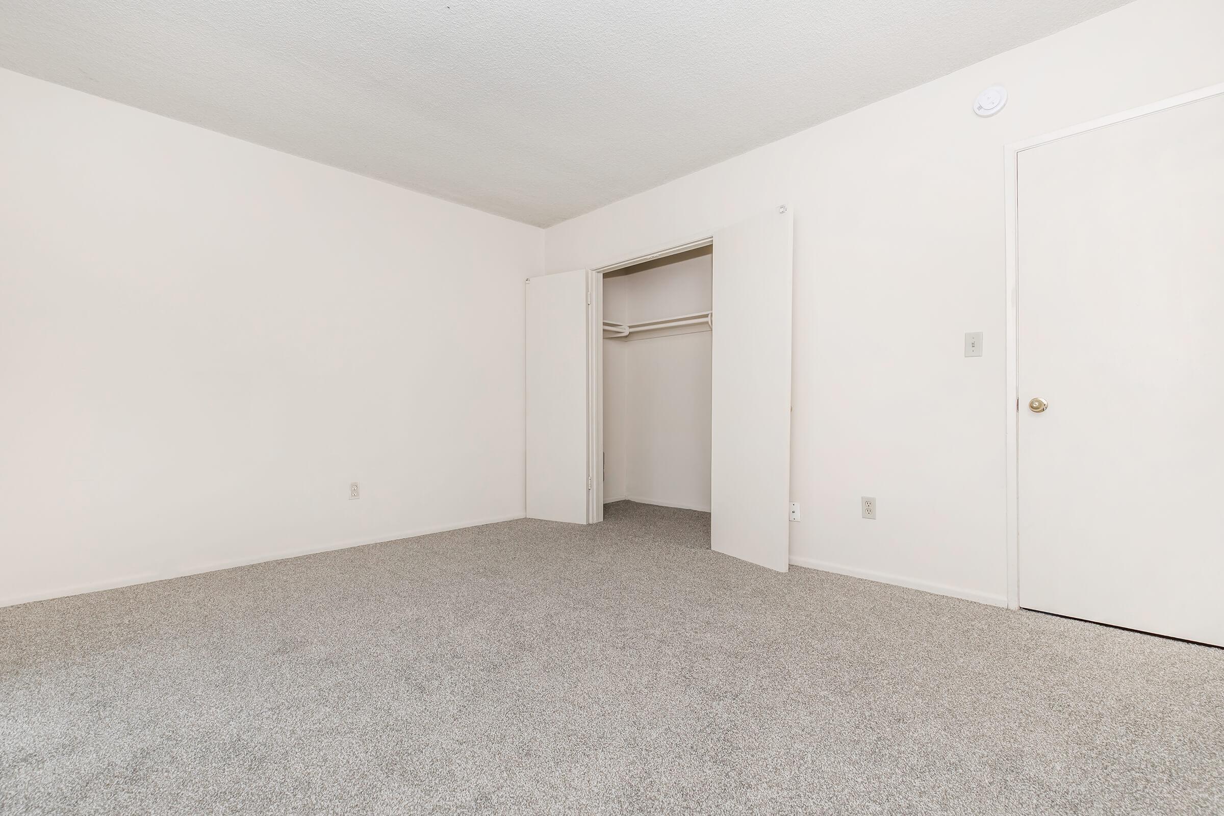 A vacant room with light-colored walls and gray carpet. One wall features an open closet with sliding doors, while another wall has a closed door. The room is spacious and well-lit, conveying a minimalist and neutral aesthetic.