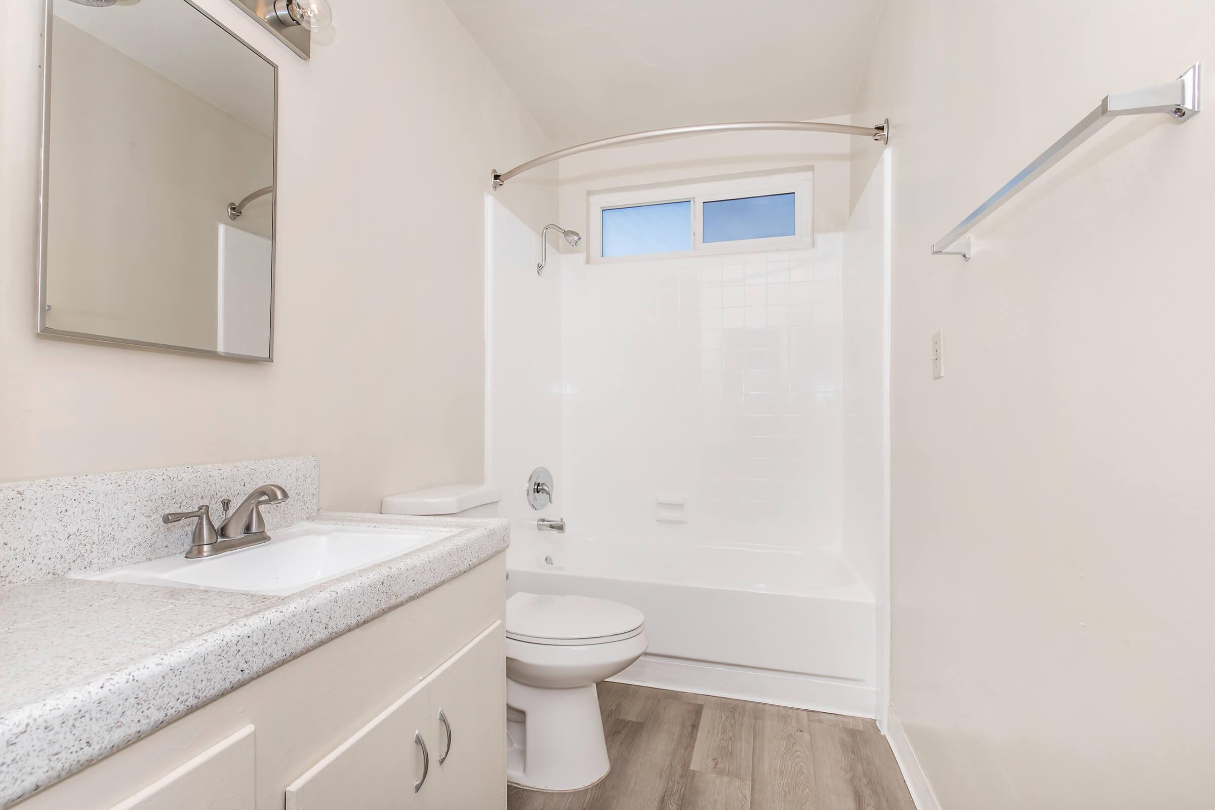 A clean and simple bathroom featuring a white bathtub with a shower, a modern sink with a faucet, a large mirror above the sink, and a towel rack. The walls are painted in a light color, and there is natural light coming through a window. The flooring is light-colored laminate.