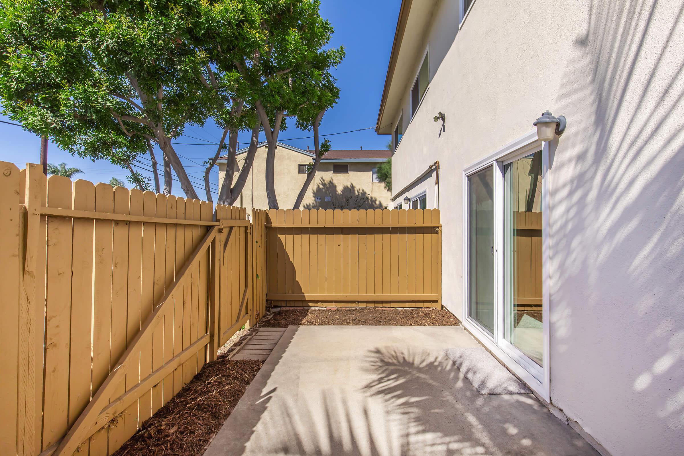 A small, enclosed outdoor patio area with a light-colored concrete floor, surrounded by a tan wooden fence. There are a few trees providing shade, and a clear blue sky overhead. The area appears well-maintained and suitable for relaxation or outdoor seating.