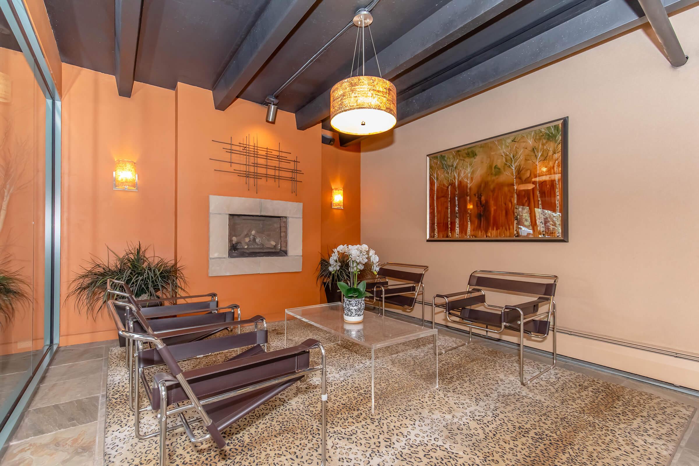 A cozy lounge area featuring sleek metal chairs around a glass table, a decorative fireplace, and a large artwork on the wall. The room has warm orange walls and a patterned rug, with potted plants adding a touch of greenery. A modern pendant light hangs from the ceiling.