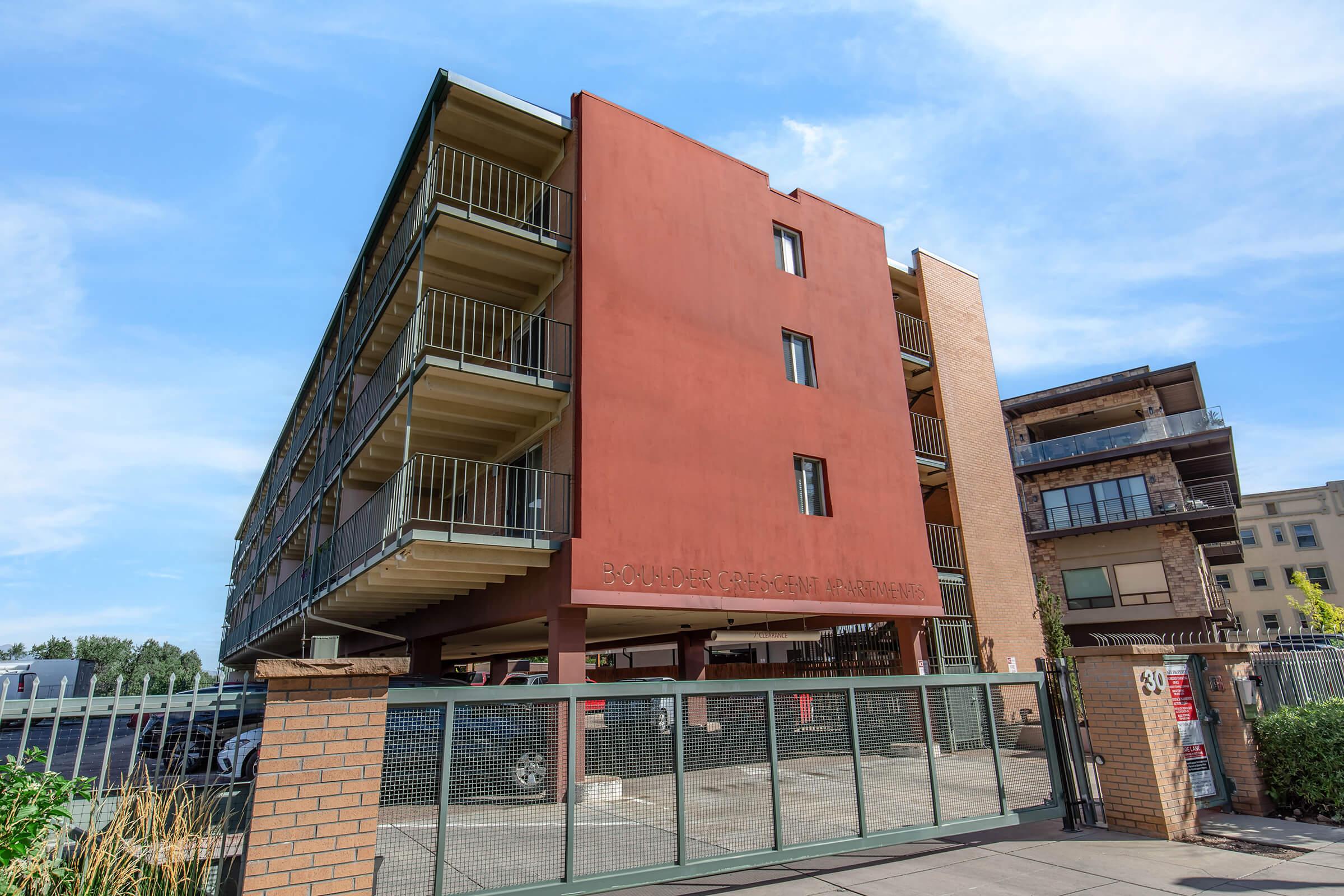 A modern multi-story residential building with a reddish exterior and green balconies. The ground level features a gated entrance. Surrounding landscaping includes grass and shrubs, and other buildings can be seen in the background under a clear blue sky.