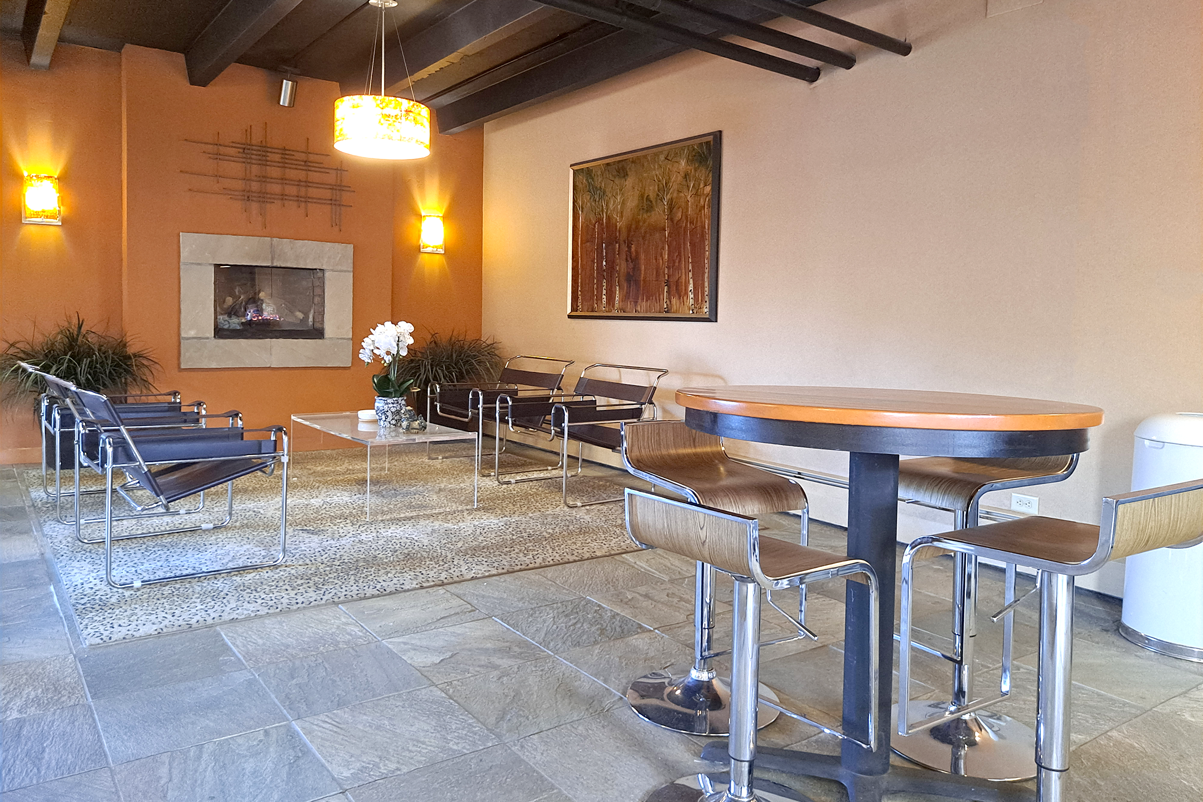 A modern lounge area featuring a cozy fireplace, comfortable seating with metal chairs and a table, and a decorative painting on the wall. The flooring is stone tiles, and there are plants for added greenery.