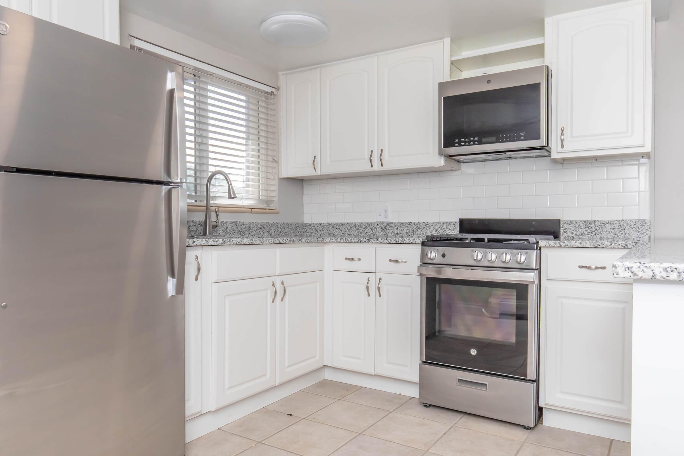A modern kitchen featuring white cabinetry, a stainless steel oven with a microwave above, a silver refrigerator, and a granite countertop. Light-colored tiles cover the floor, and there are window blinds allowing natural light into the space.