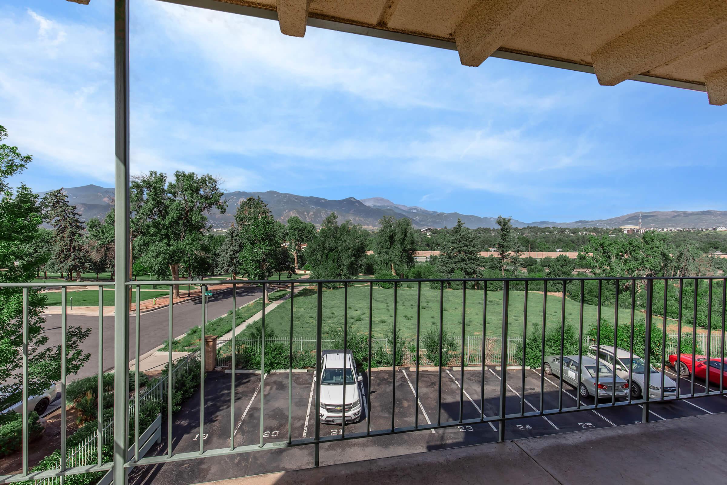 View from a balcony overlooking a parking area with several cars. In the background, there are trees and mountains, with a clear blue sky above. The scene suggests a serene, natural environment.
