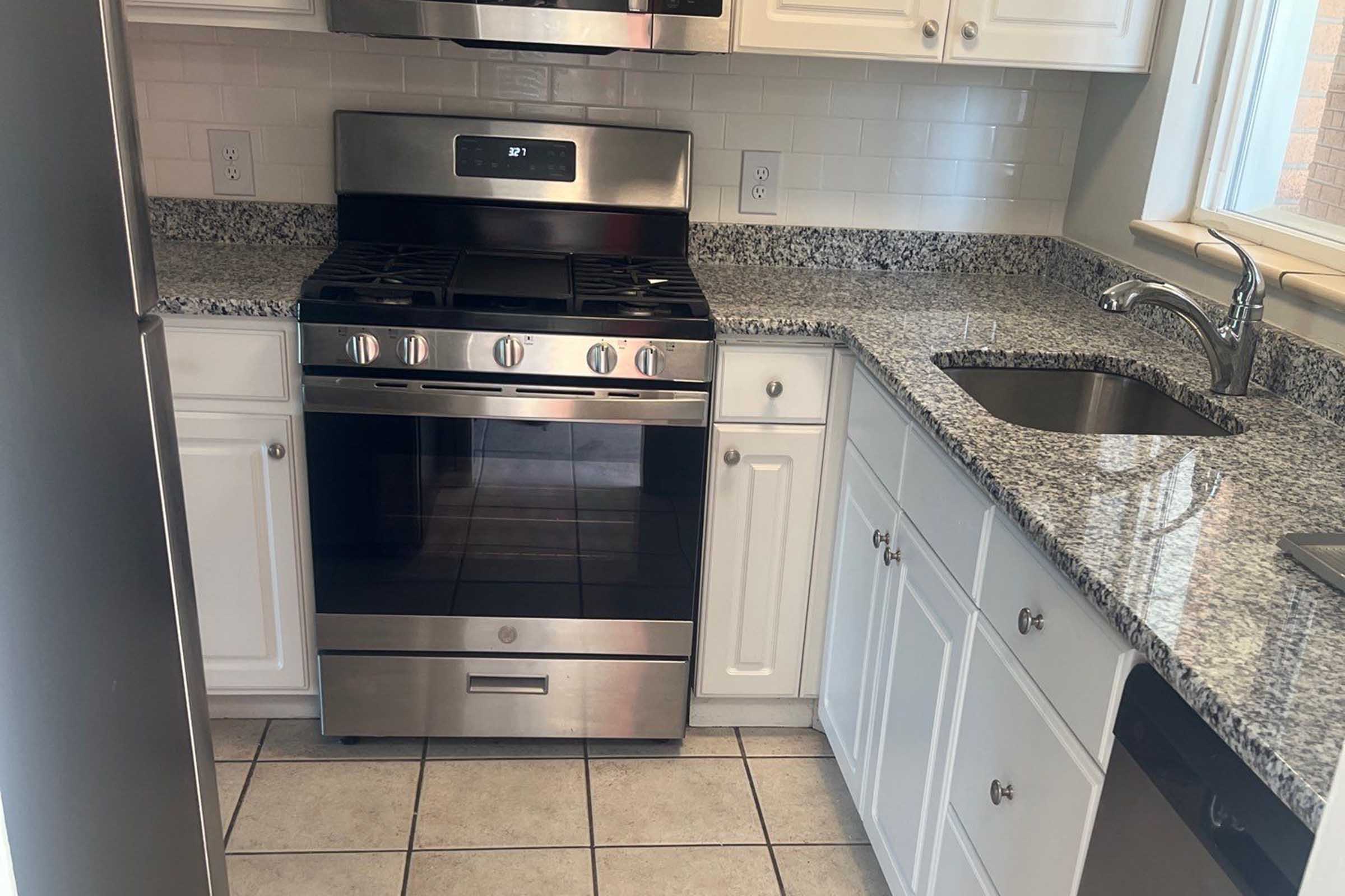 A modern kitchen featuring stainless steel appliances, including a gas range and an overhead microwave. The countertops are made of granite, complemented by white cabinetry. A stainless steel sink is located near a window, and the flooring consists of square tiles.