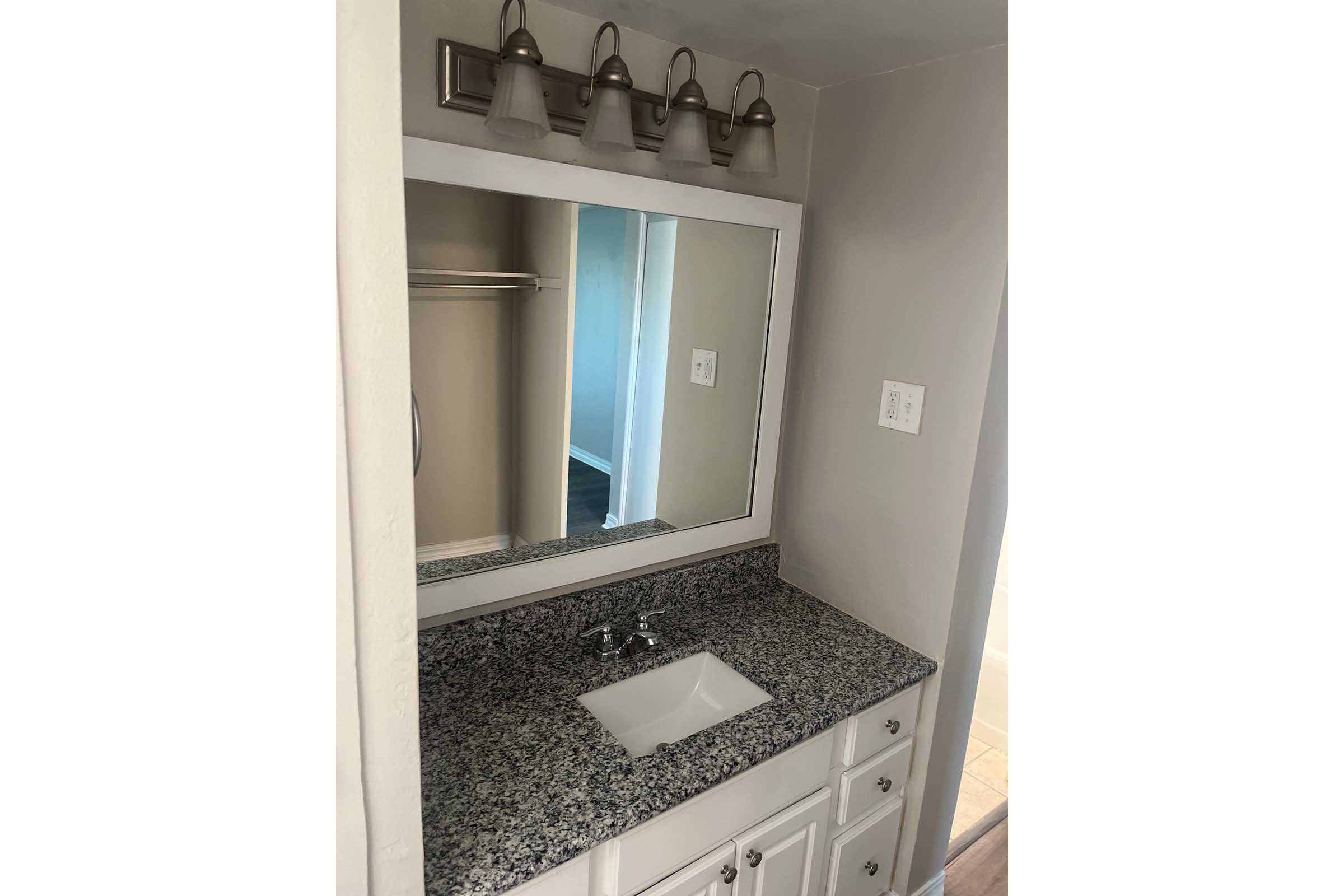 A modern bathroom vanity with a large mirror, granite countertop, and a white sink. The area is well-lit by four wall-mounted light fixtures above the mirror. The space features neutral beige walls and a doorway leading to another room in the background.