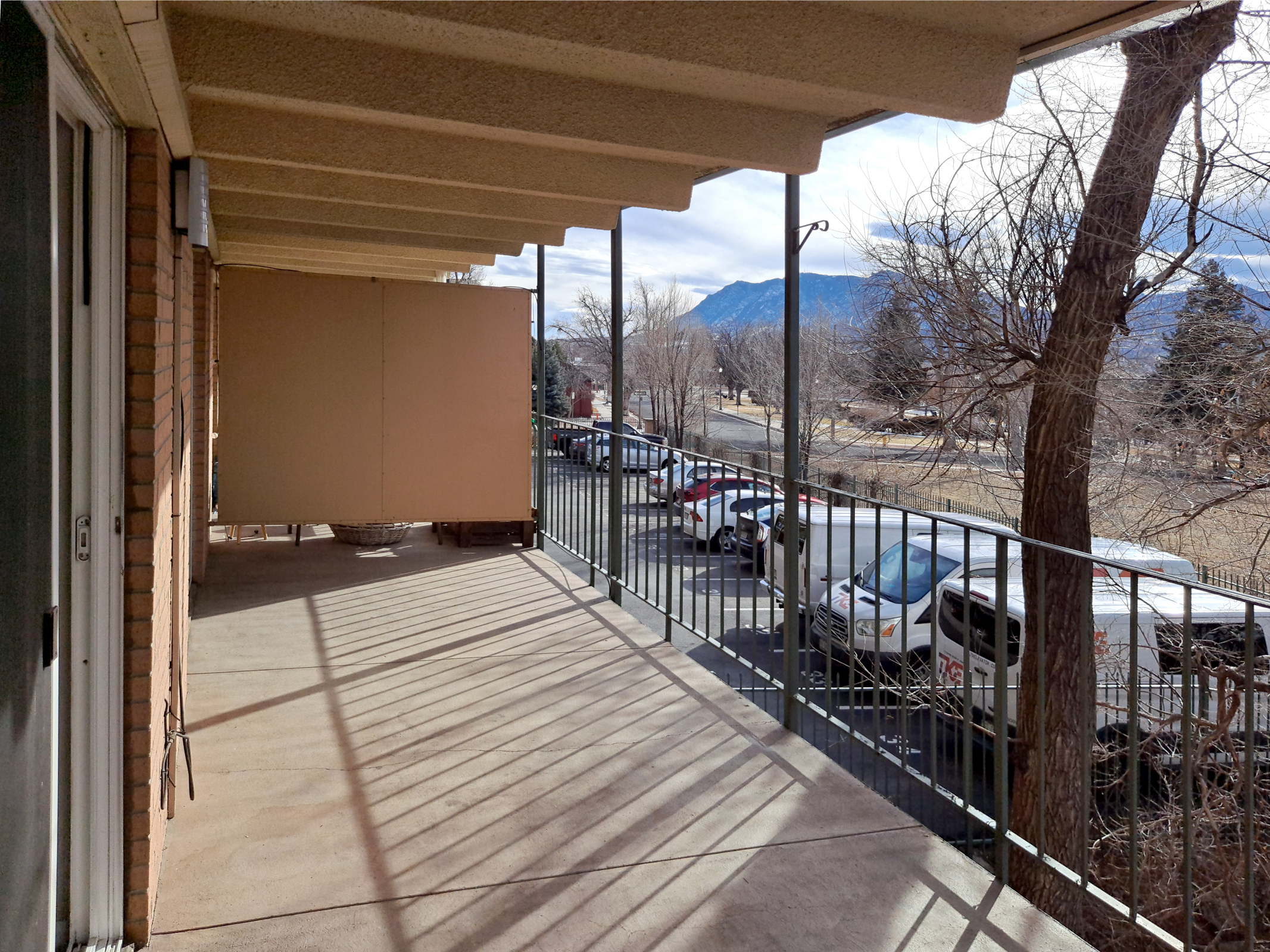 A spacious balcony with a railing, displaying shadows cast by the overhang. In the background, parked cars line a street, with trees and mountains visible in the distance under a cloudy sky. The scene conveys a quiet, suburban atmosphere.