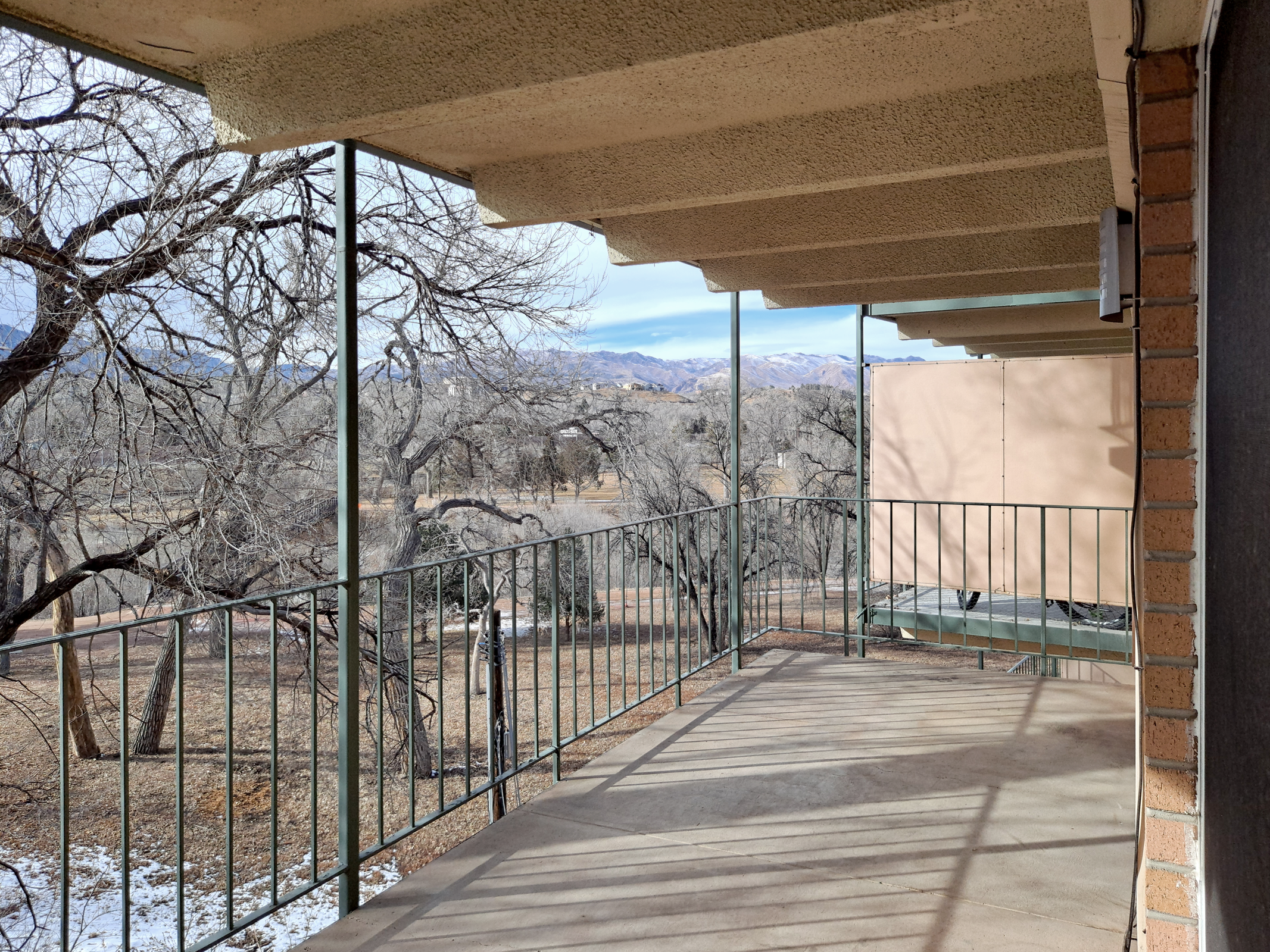 View from a covered balcony featuring a railing, overlooking a landscape with bare trees and mountains in the distance. The ground is bare with some patches of grass, suggesting early spring or fall. Natural light illuminates the scene, creating a serene atmosphere.