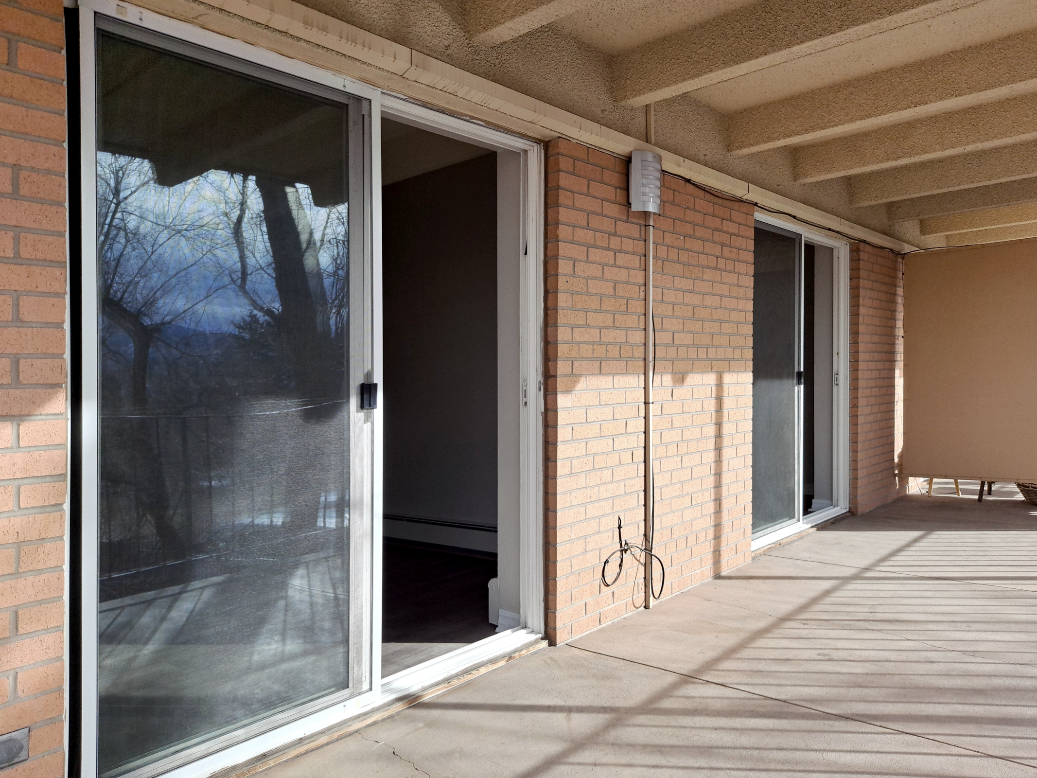 A spacious balcony with two sliding glass doors leading indoors. The walls are brick, and there are shadows cast on the floor from nearby trees. The light creates a bright atmosphere, and there is a utility cable visible along the wall.