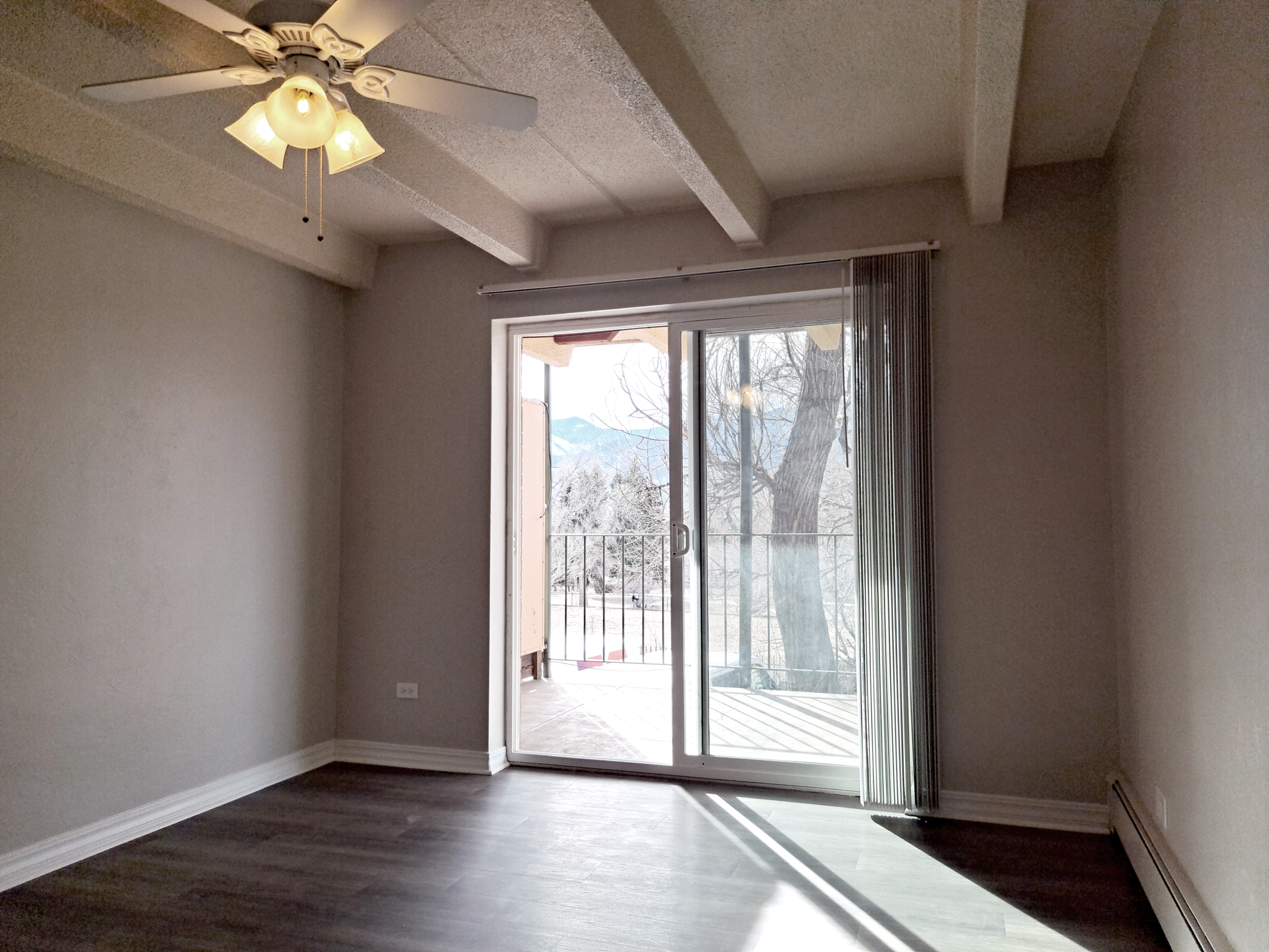 A well-lit room with a ceiling fan, featuring a sliding glass door that leads to a balcony. The room has light gray walls and dark wood-like flooring, with visible mountain scenery outside. Natural light streams in, creating a warm and inviting atmosphere.