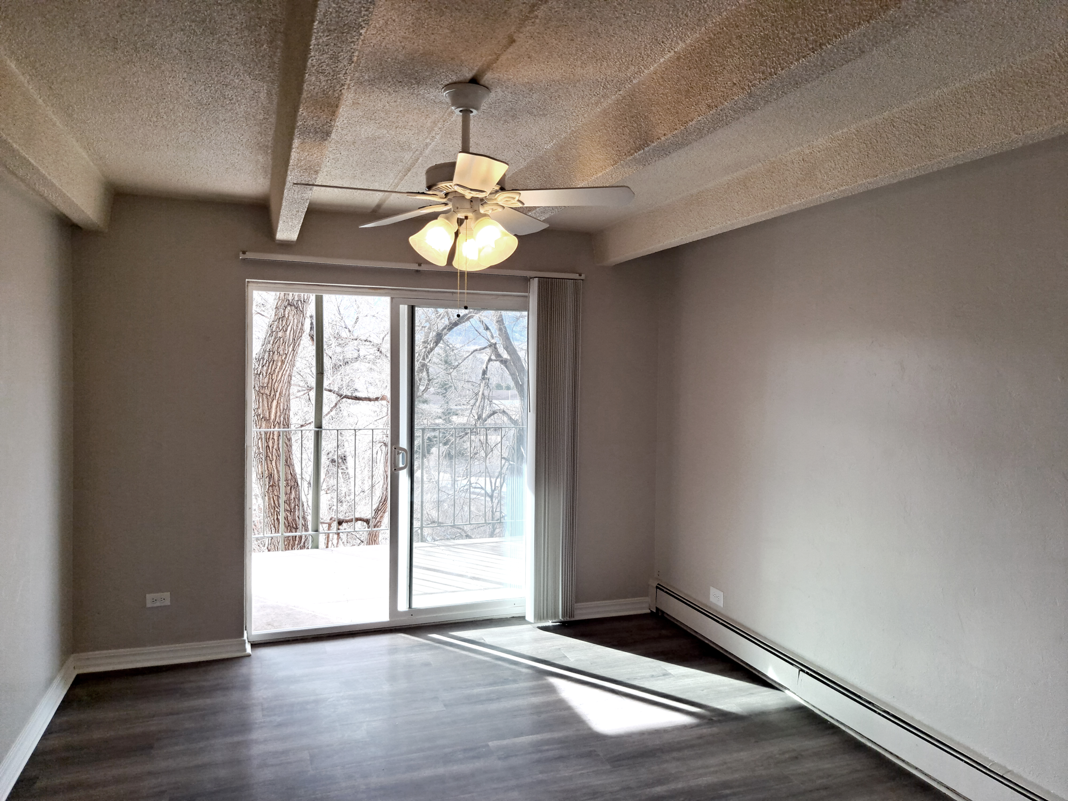 A bright, empty living room with light gray walls and a ceiling fan featuring four lights. Large sliding glass doors open to a balcony, allowing natural light to fill the space. The floor is dark-colored, and there is a sense of spaciousness in the area.