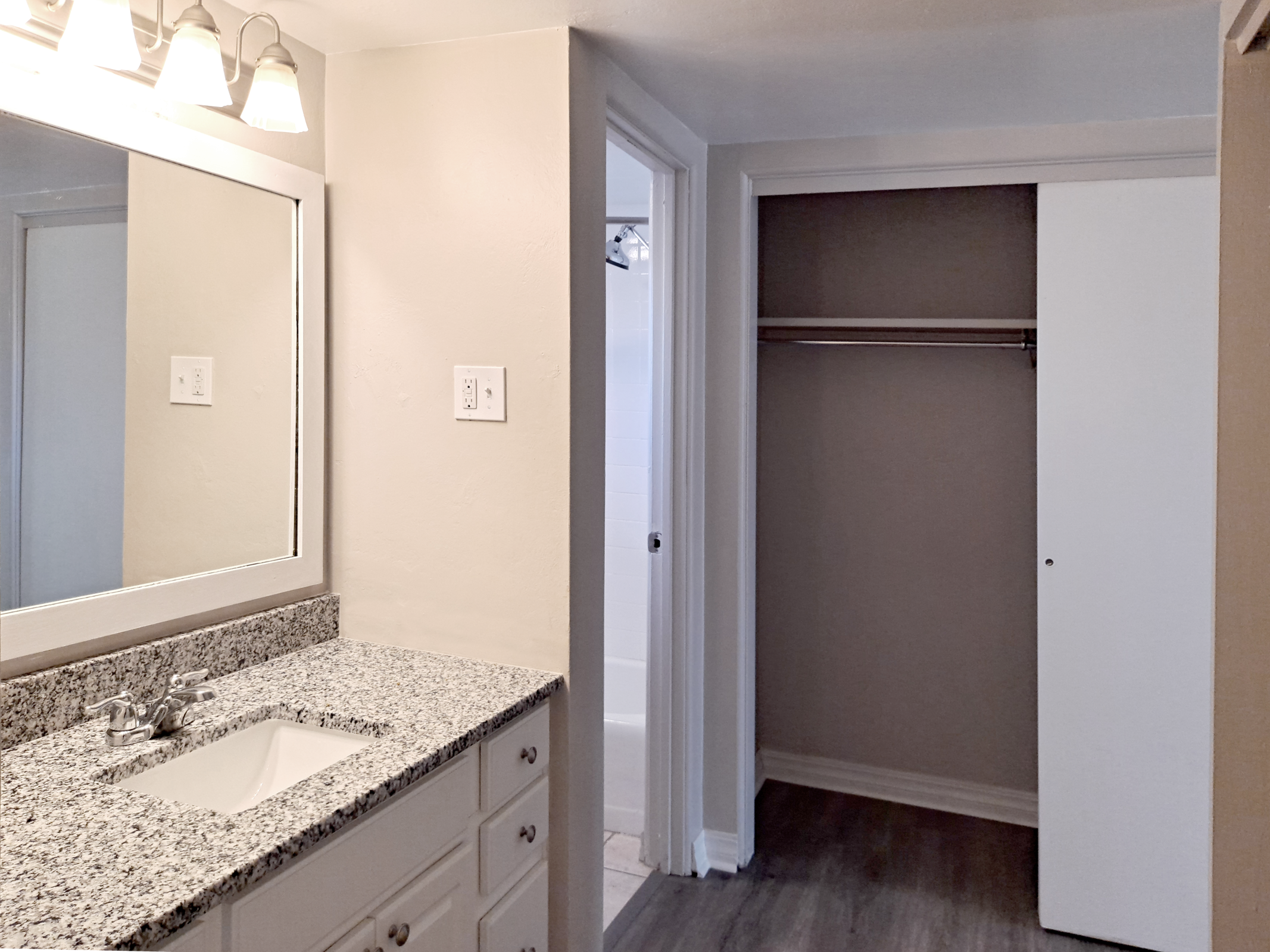 A bright and modern bathroom featuring a vanity with a granite countertop and a mirror above it. The bathroom includes a doorway leading to a tub area and an adjacent closet with a sliding door. The walls are painted in light colors, contributing to an airy and spacious feel.