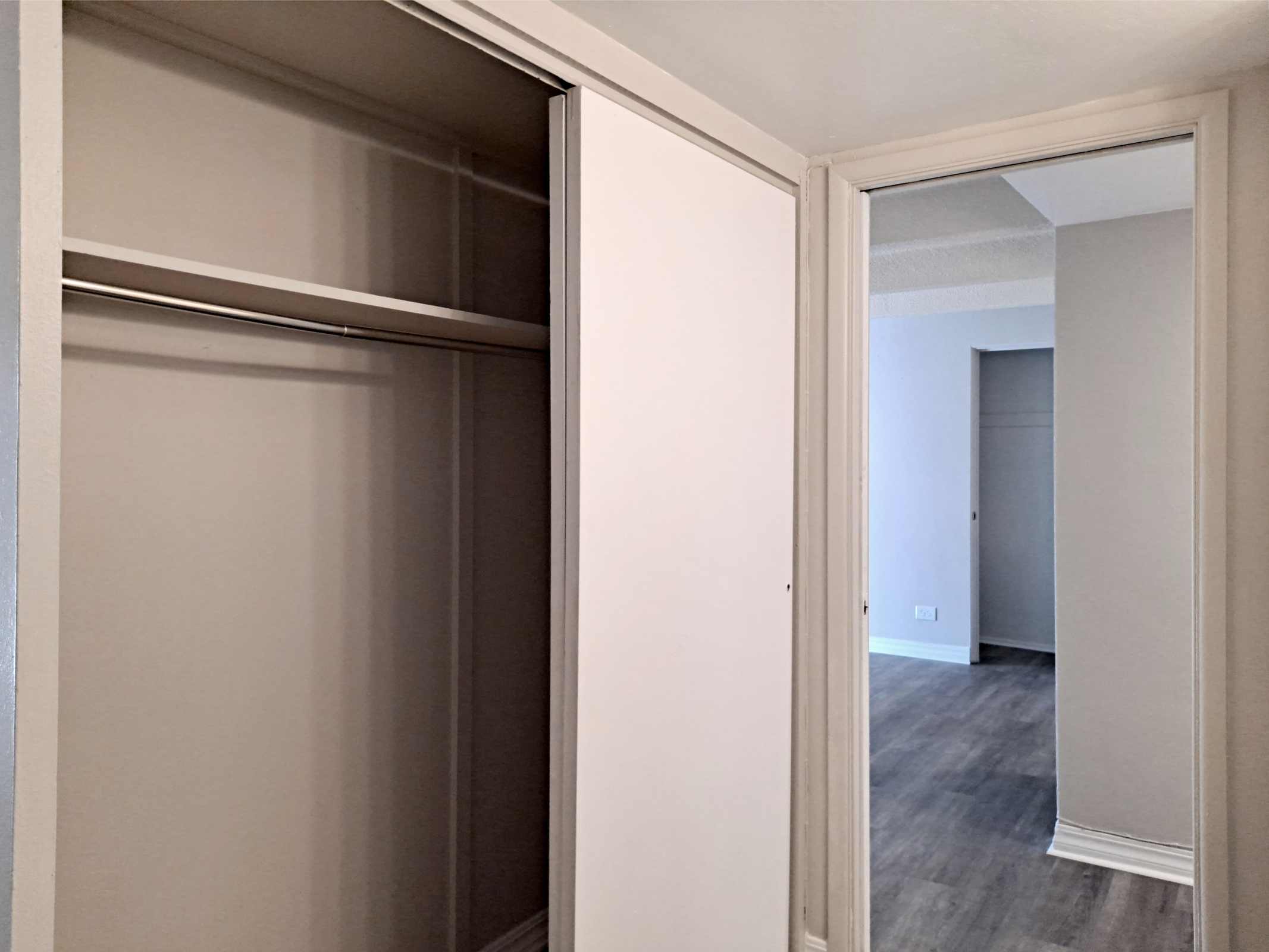 An empty closet with sliding doors and a mirror on the right side. The walls are painted light gray, and the wooden floor is visible in the background, leading to a well-lit room with minimal furniture. The interior has a clean and simple design.