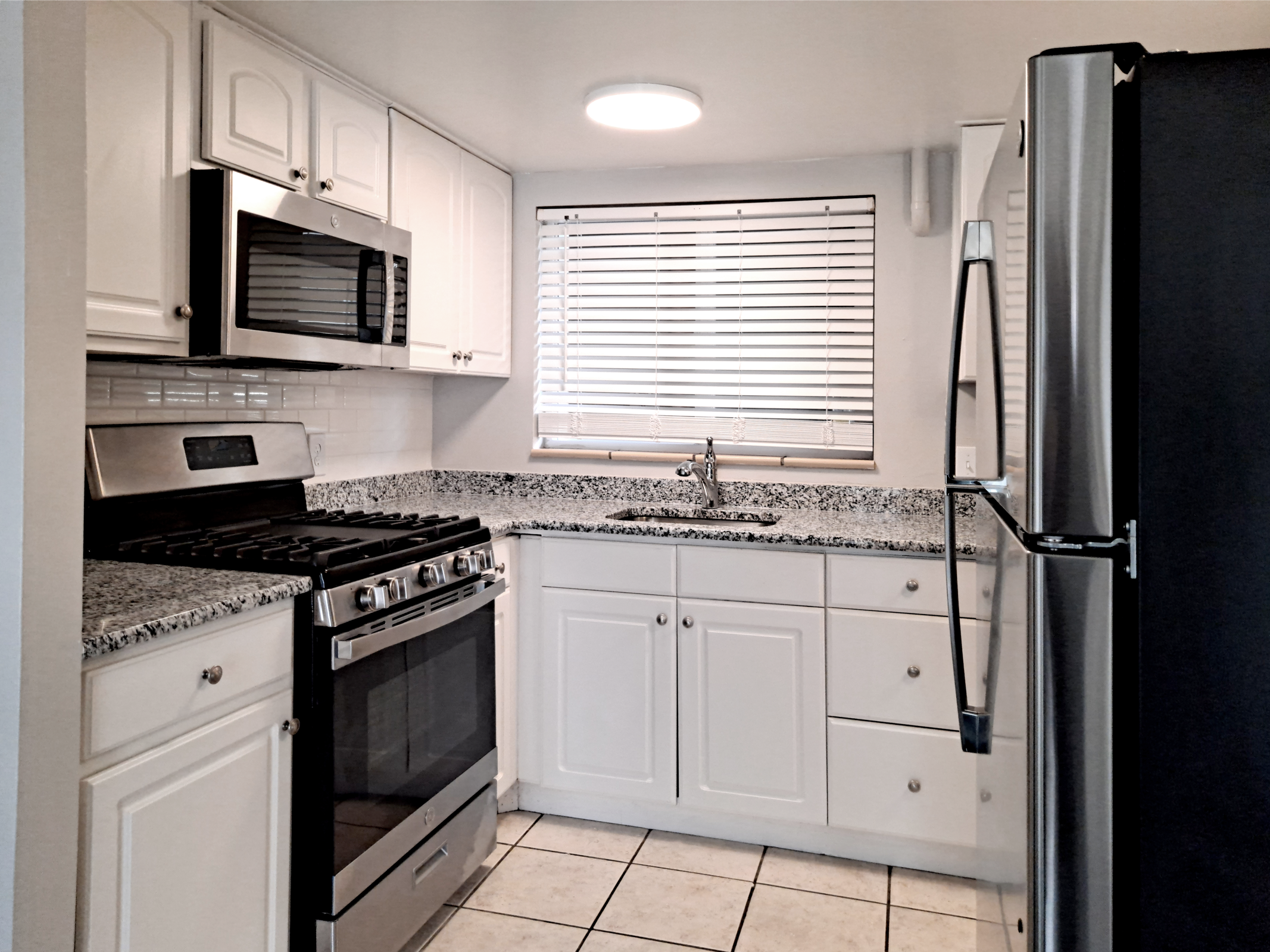 Modern kitchen featuring white cabinetry, a stainless steel refrigerator, and a gas stove with an oven. The countertops are made of gray and white speckled granite. There's a window with blinds above the sink, providing natural light. The floor is tiled in a simple layout.