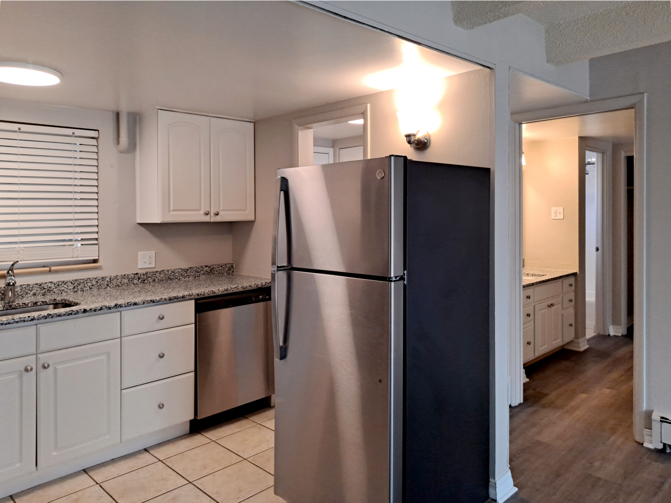 A modern kitchen featuring stainless steel appliances, including a refrigerator and dishwasher, light-colored cabinets with granite countertops, and a window with blinds. The image shows an open layout with a hallway leading to a bathroom area, well-lit by overhead lights.