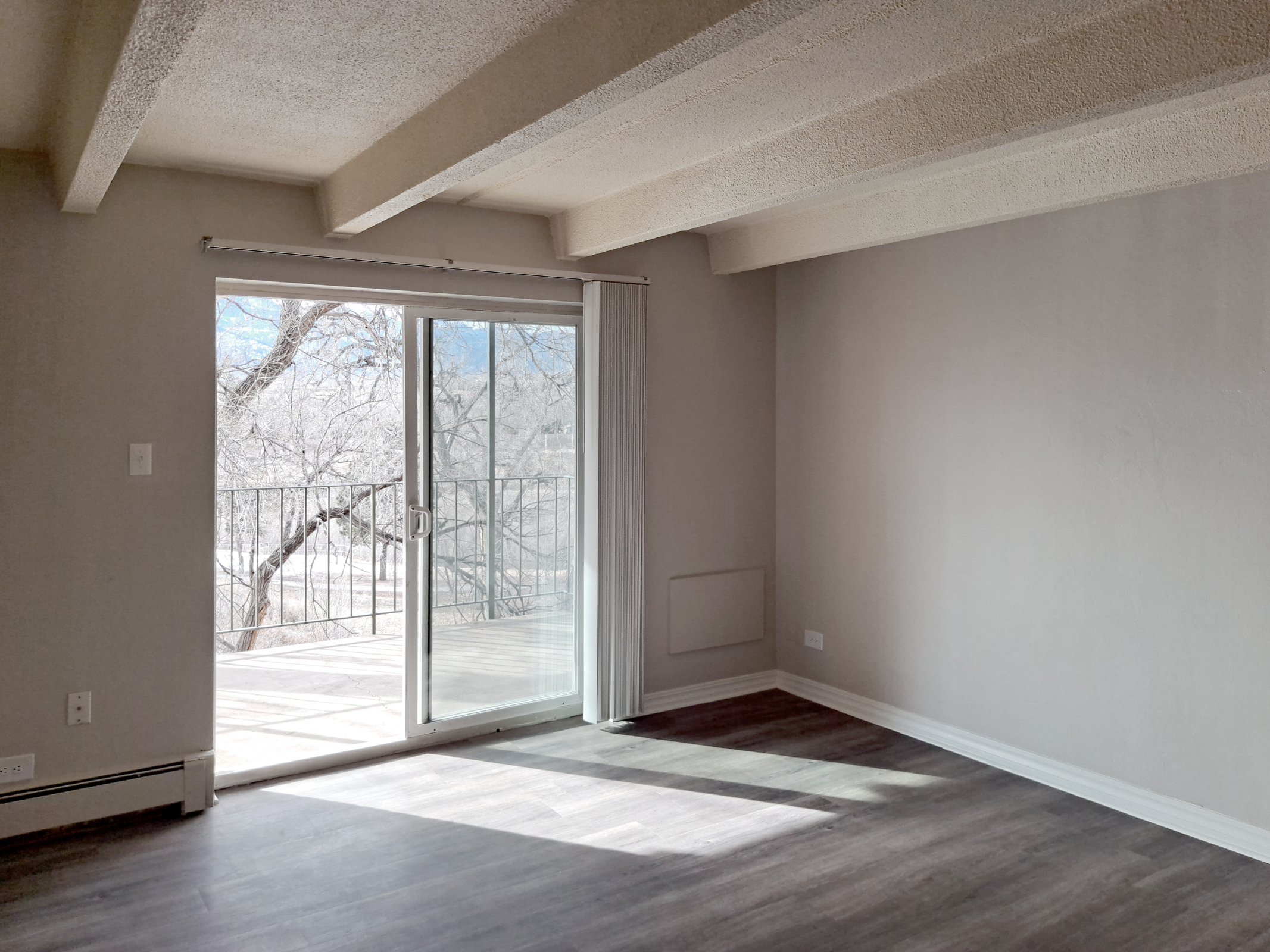 A spacious, empty room with light gray walls and a wooden floor. It features a large sliding glass door that leads to a balcony, allowing natural light to fill the space. The ceiling has exposed beams, and a corner shows a bit of the outdoor view, indicating it's a bright, inviting area.