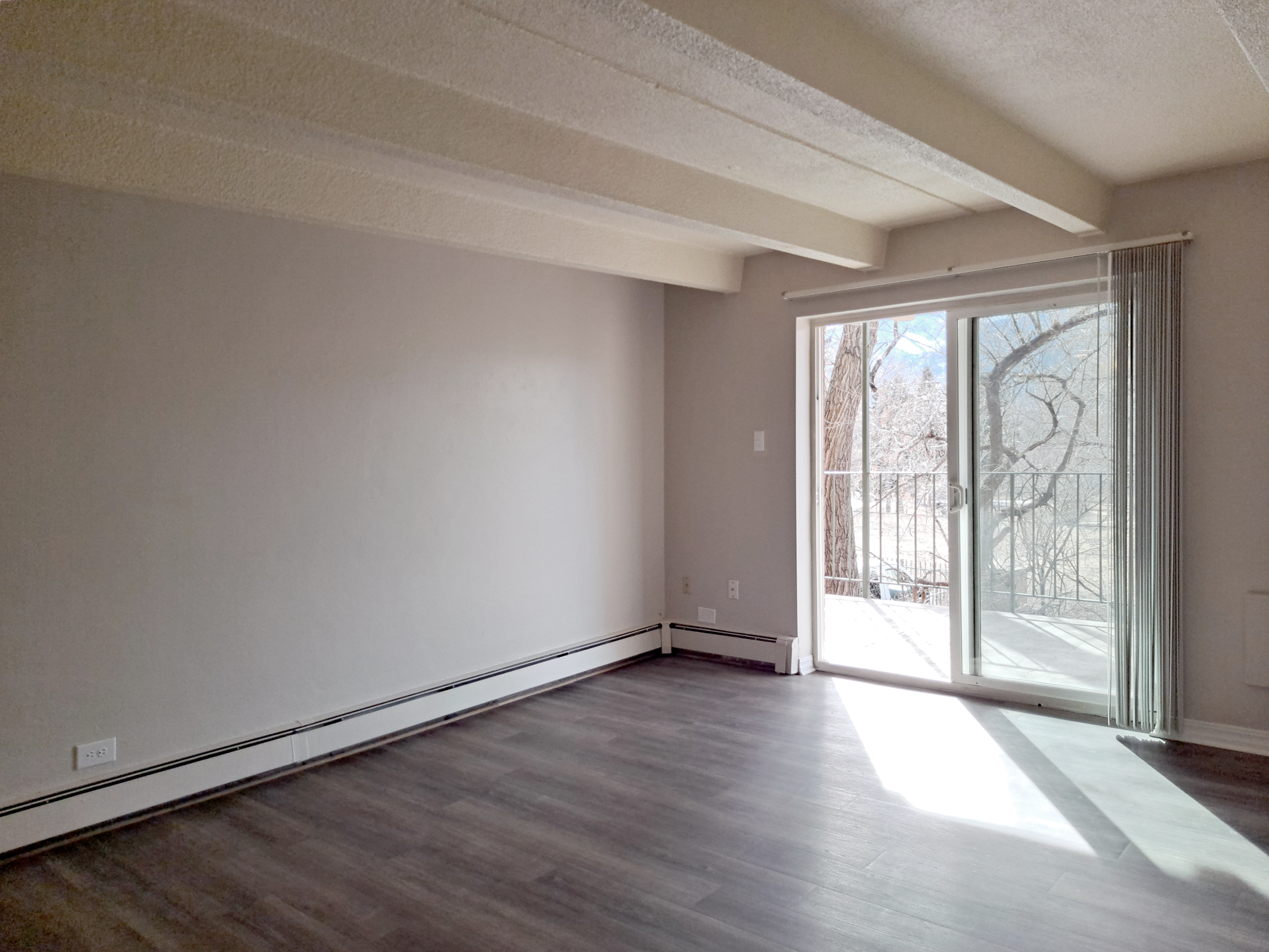 A spacious, empty room with light gray walls and a large sliding glass door leading to a balcony. The floor is covered in dark wood laminate. Natural light fills the space, highlighting the minimalist design and exposed ceiling beams. No furniture is present, creating a clean, open atmosphere.