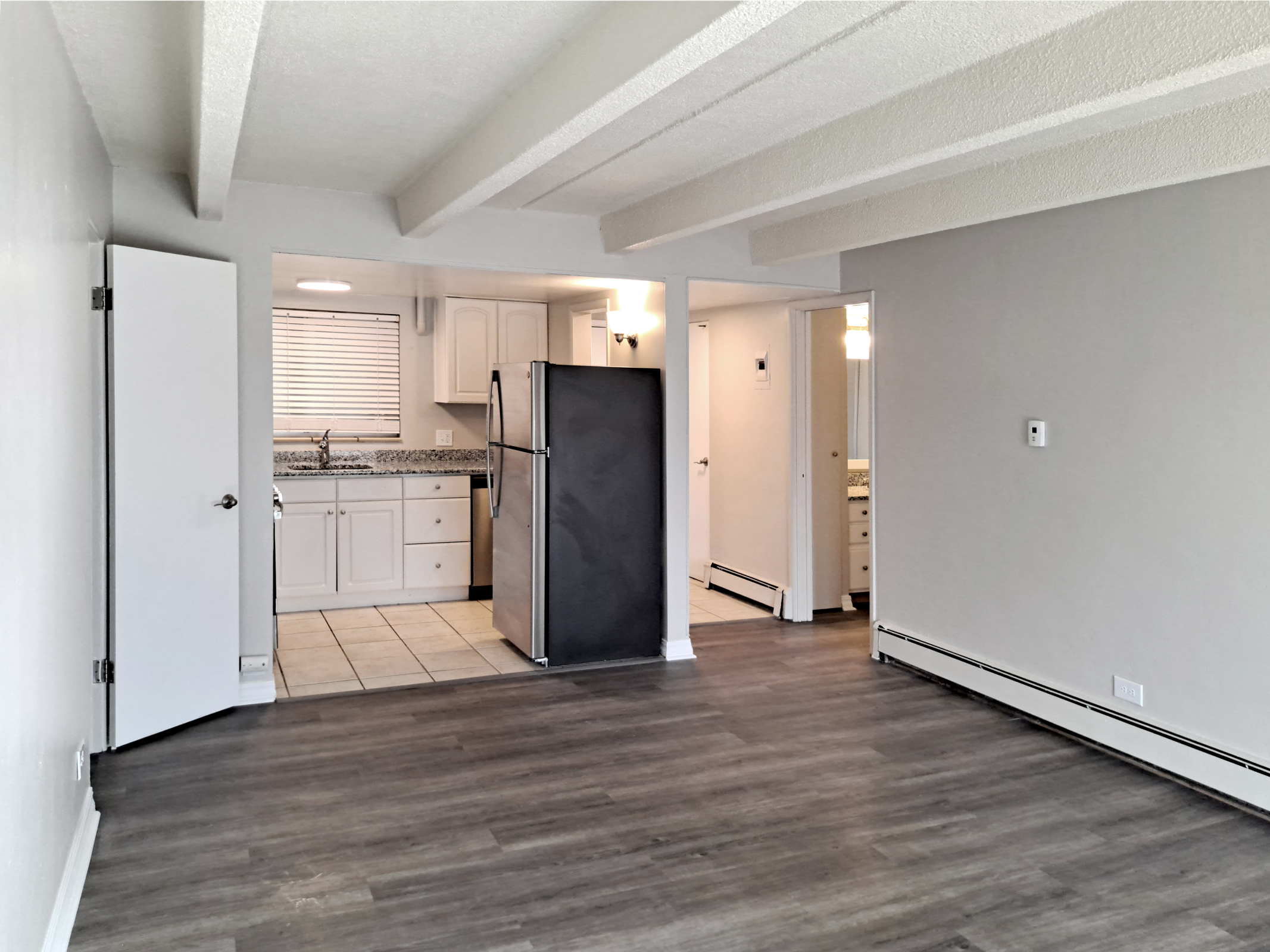 An interior view of a modern living space featuring an open floor plan. The room includes a kitchen area with white cabinets and a granite countertop, a stainless steel refrigerator, and a door leading to a bathroom. The floor is wooden, and the walls are painted in a light neutral color.