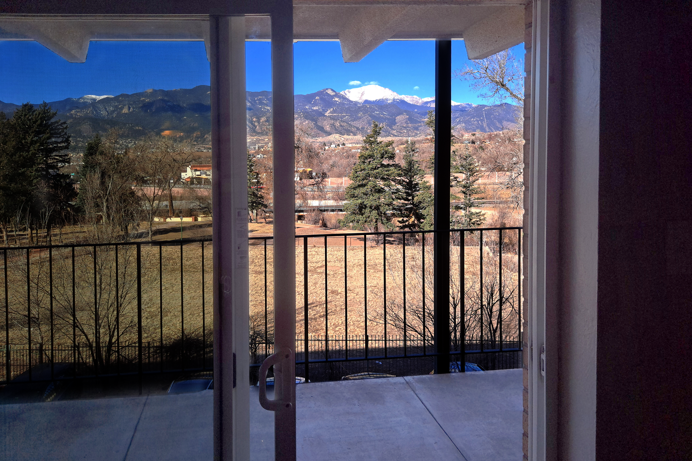 A view from an open door or window showing a balcony overlooking a landscape. In the foreground, there are trees and a grassy area, while in the background, snow-capped mountains rise against a clear blue sky. The scene captures a tranquil outdoor setting with a blend of natural beauty.