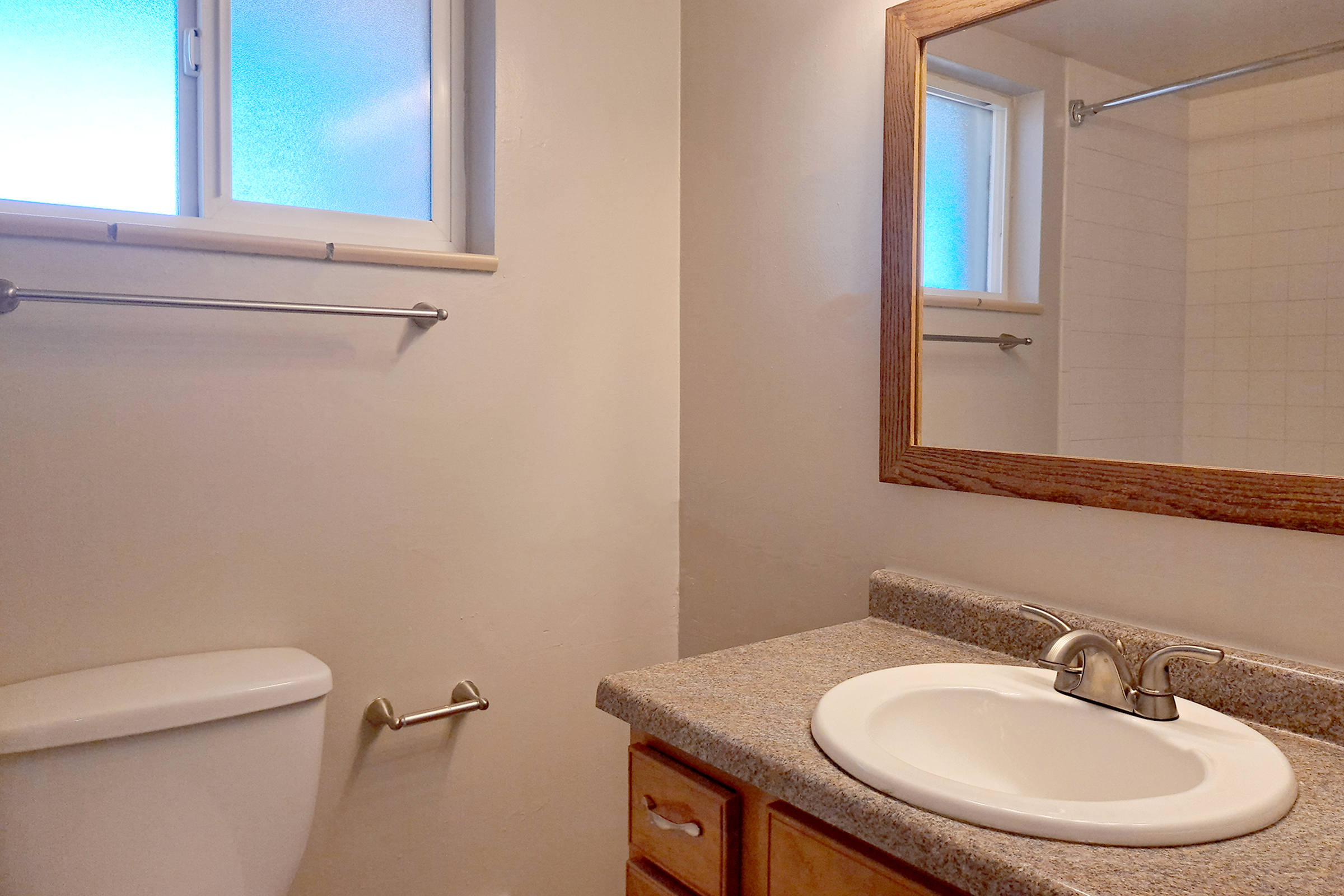 A clean bathroom featuring a toilet, a sink with a simple faucet, and a mirror framed in wood. The walls are painted a light color, and there are two windows allowing natural light to enter. A countertop made of speckled gray material complements the wooden cabinetry.