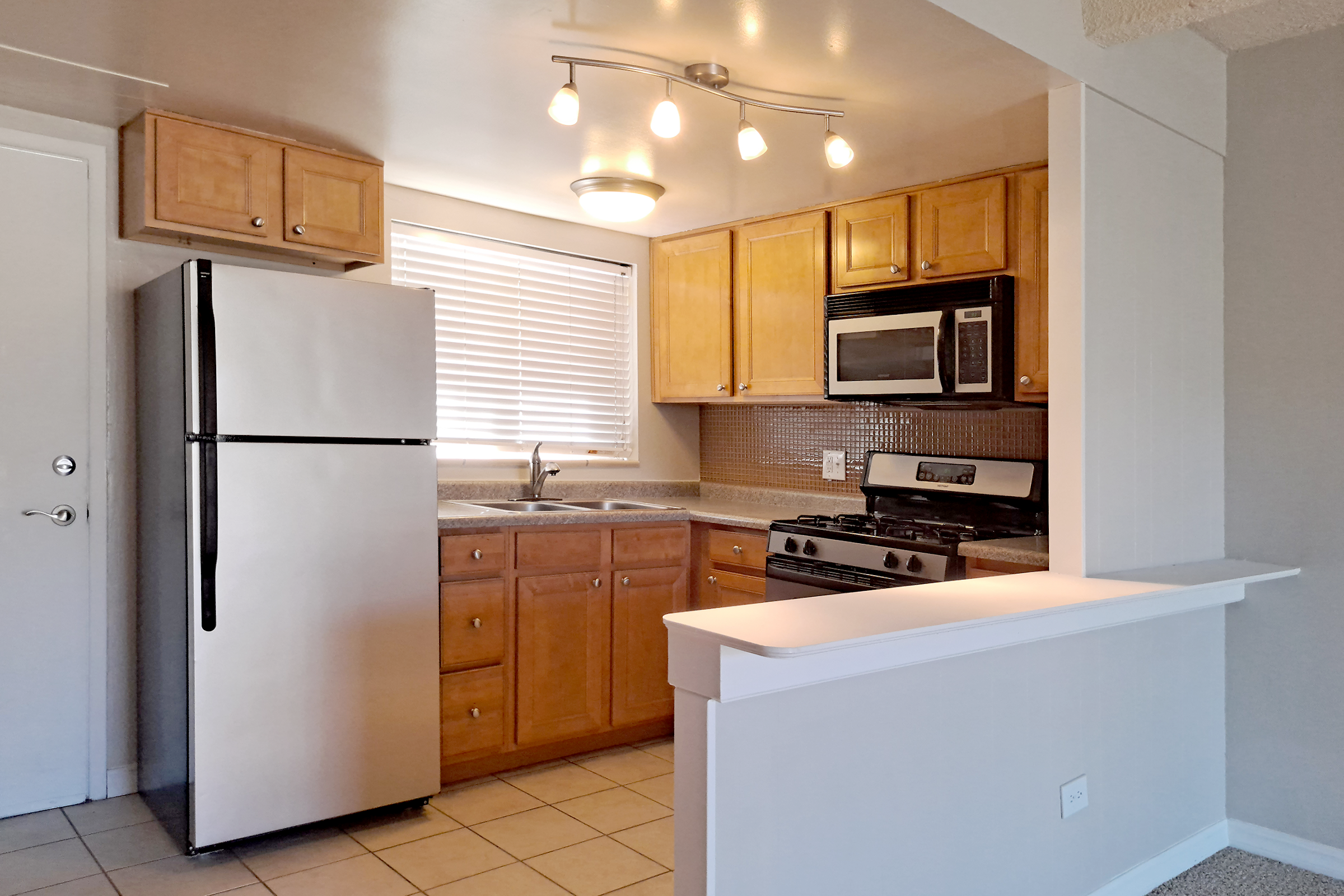 A modern kitchen featuring light wood cabinets, a stainless steel refrigerator, a gas stove, and a microwave. The countertop has a sink with a window above it, allowing natural light. The space includes pendant lighting and beige tiled flooring.