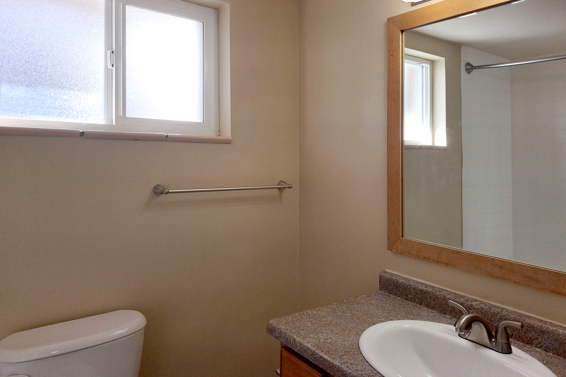 A clean, neutral-toned bathroom featuring a small window for natural light, a wall-mounted towel rack, a modern sink with a chrome faucet, and a large mirror above the vanity. The walls are pale, and the bathroom has a spacious, airy feel.